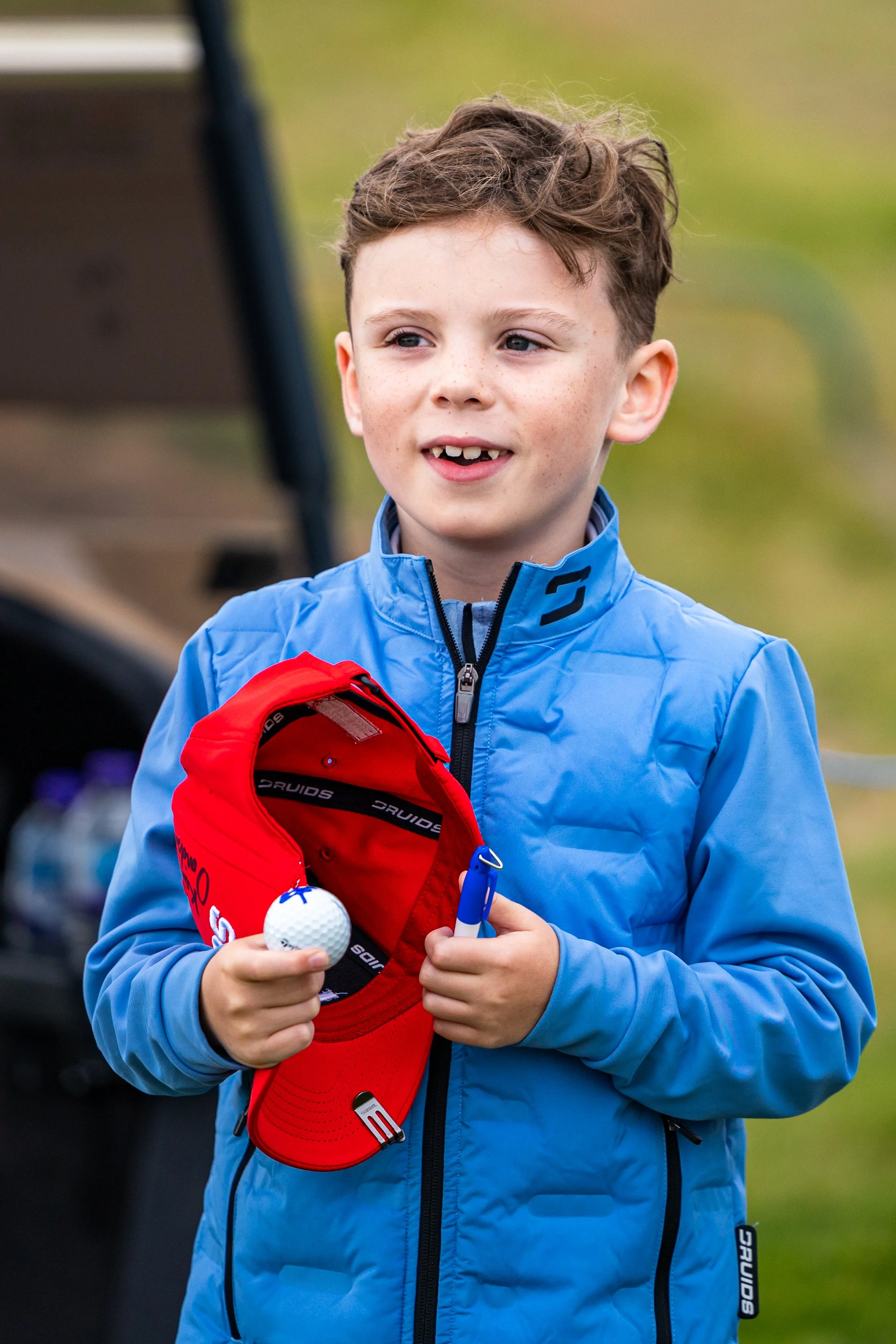 A young boy with short brown hair and freckles wearing a blue jacket holds a red golf bag, a golf ball, and a marker, standing outdoors on a golf course with a blurred background.