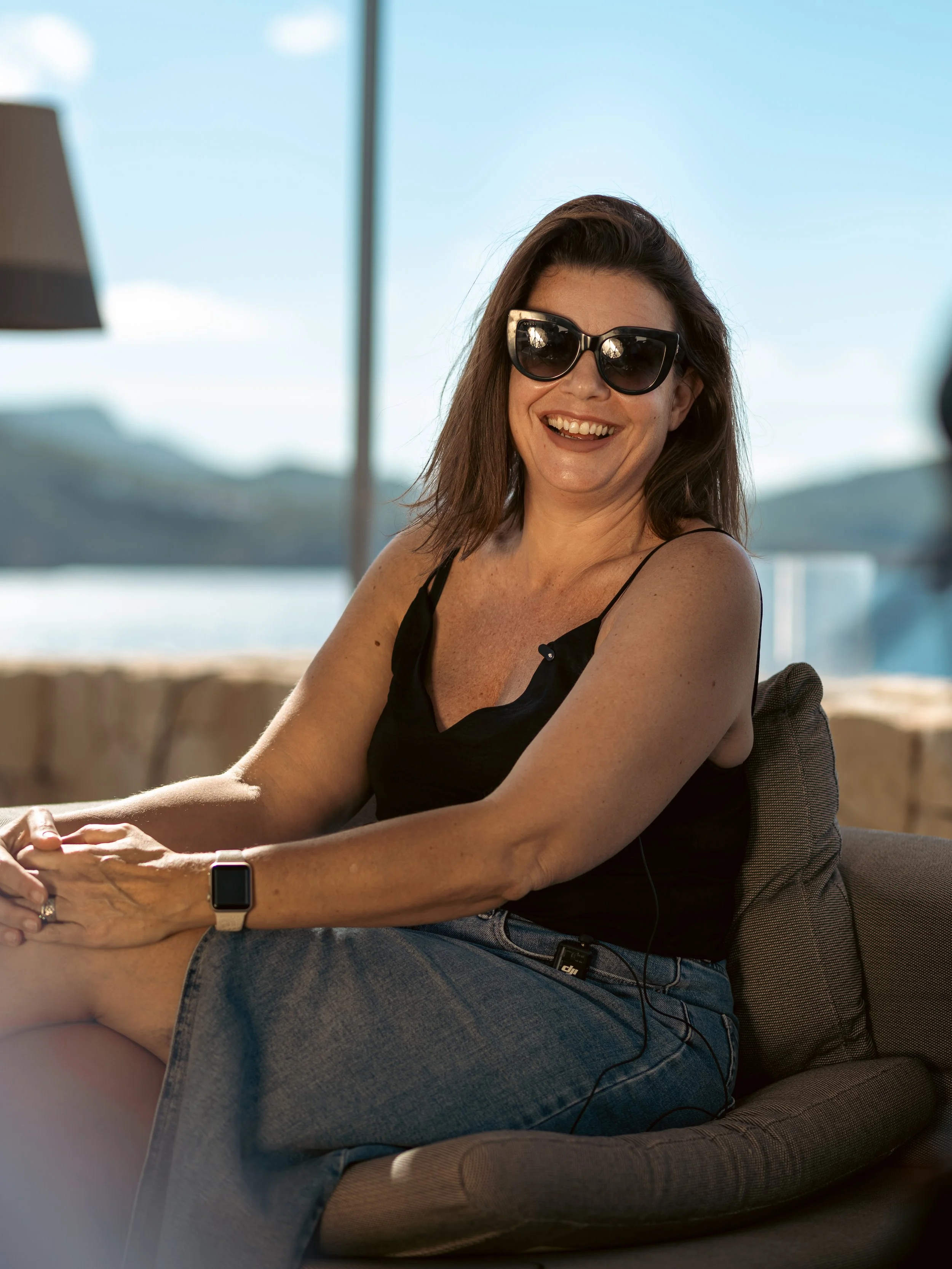 Woman with dark hair wearing sunglasses, black tank top, and watch, smiling while sitting on a gray armchair with a scenic outdoor background.
