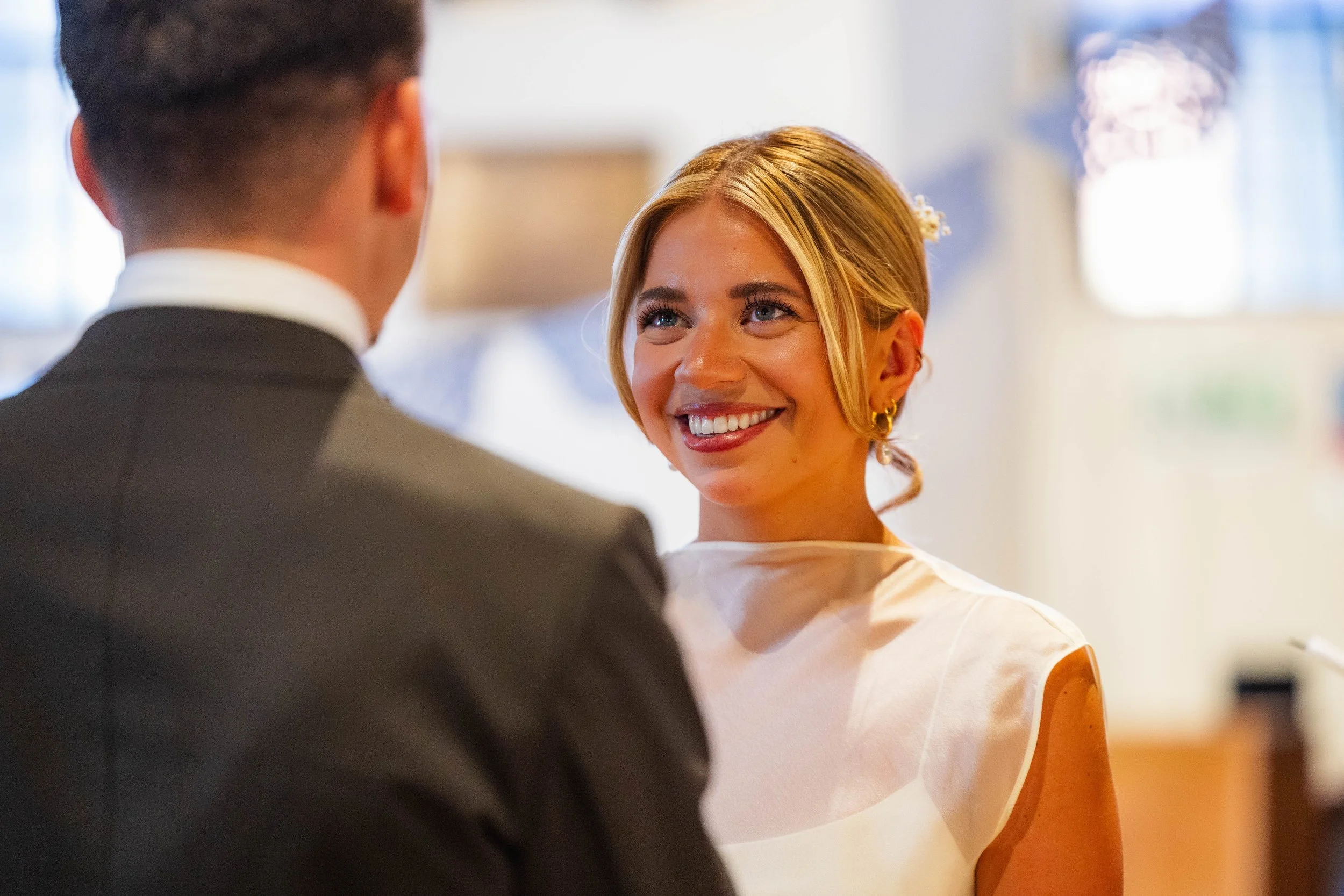A woman with blonde hair smiling and looking at a man dressed in a suit, during what appears to be a wedding or formal event.