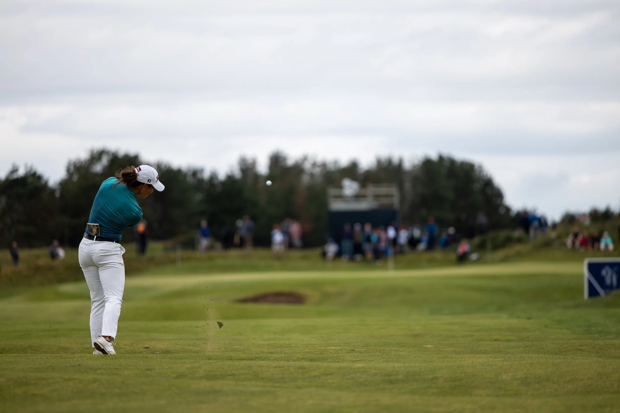 A female golfer in a teal shirt and white pants is hitting a golf ball on a golf course. She is wearing a white cap and your golf club is in motion, sending the ball into the air. In the background, spectators and trees are visible under a cloudy sky
