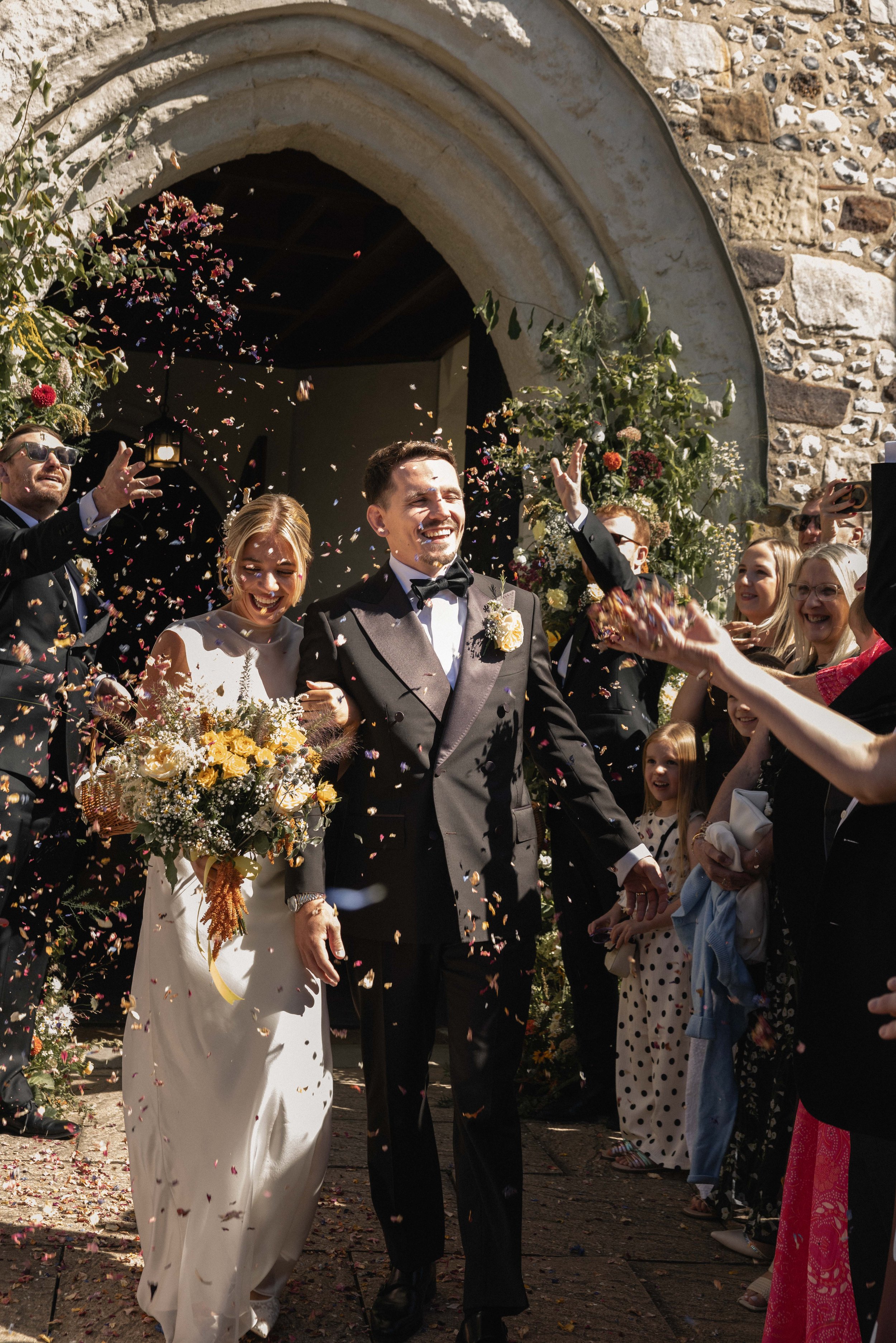 A newlywed couple is walking out of a building, smiling, as friends and family throw colorful confetti to celebrate. The bride is in a white gown holding a bouquet, and the groom is in a black tuxedo. Guests are dressed in various formal and semi-for