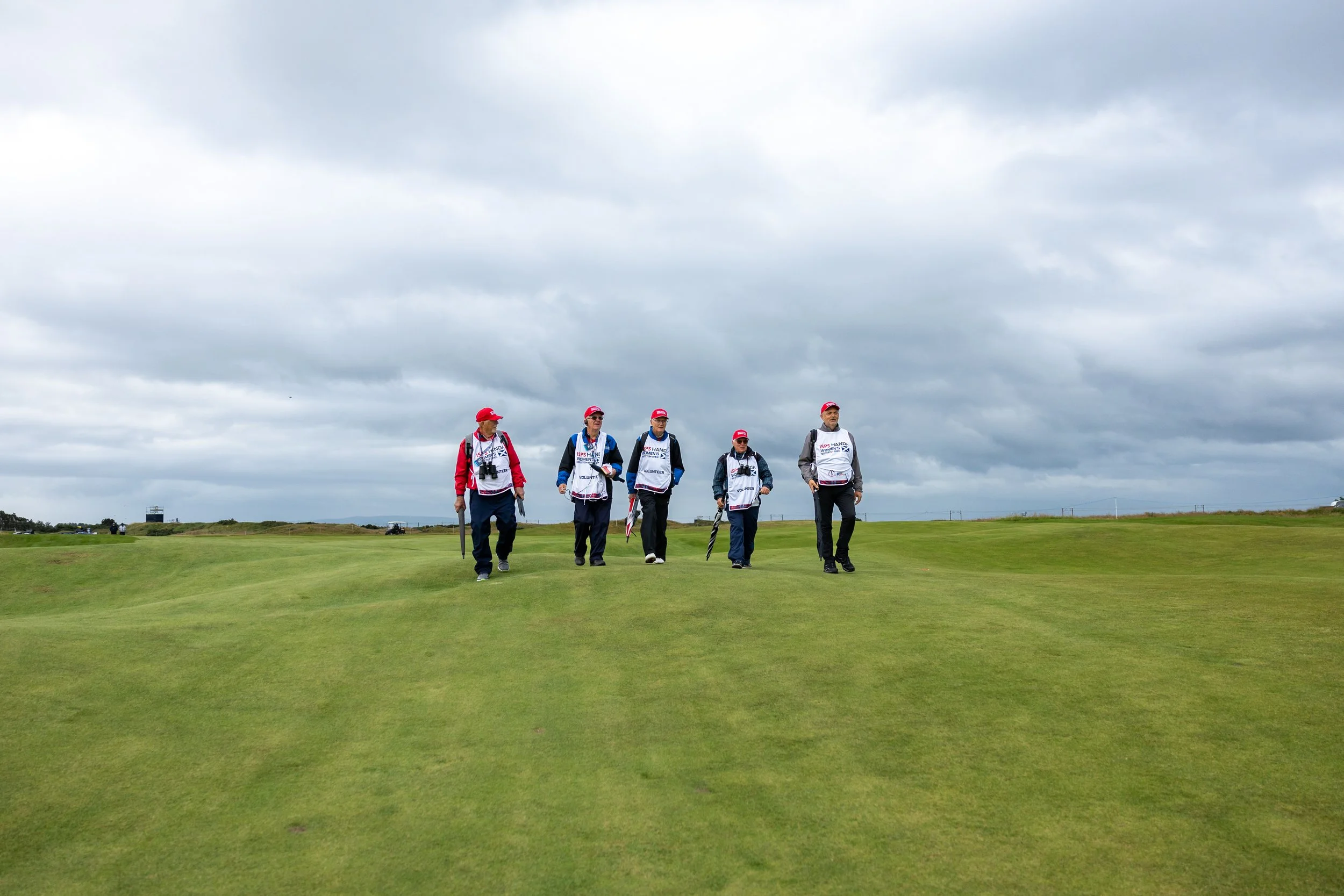 A group of six individuals walking across a golf course on a cloudy day. They are wearing matching vests and red caps, carrying umbrellas, possibly participating in a volunteer activity.