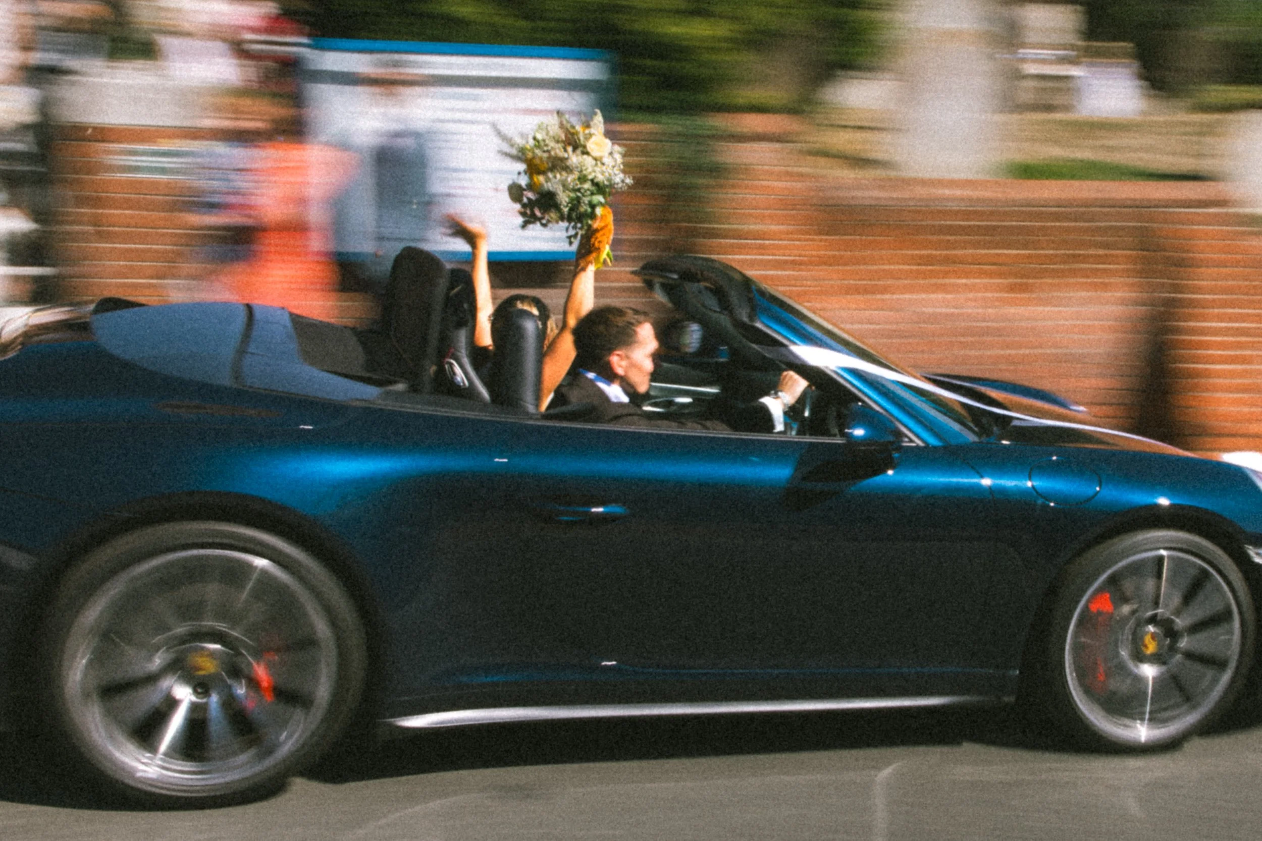 A dark blue convertible sports car driving on a road amidst a blurred background of greenery and brick walls. There are two people inside, with the woman holding a bouquet of flowers above her head.