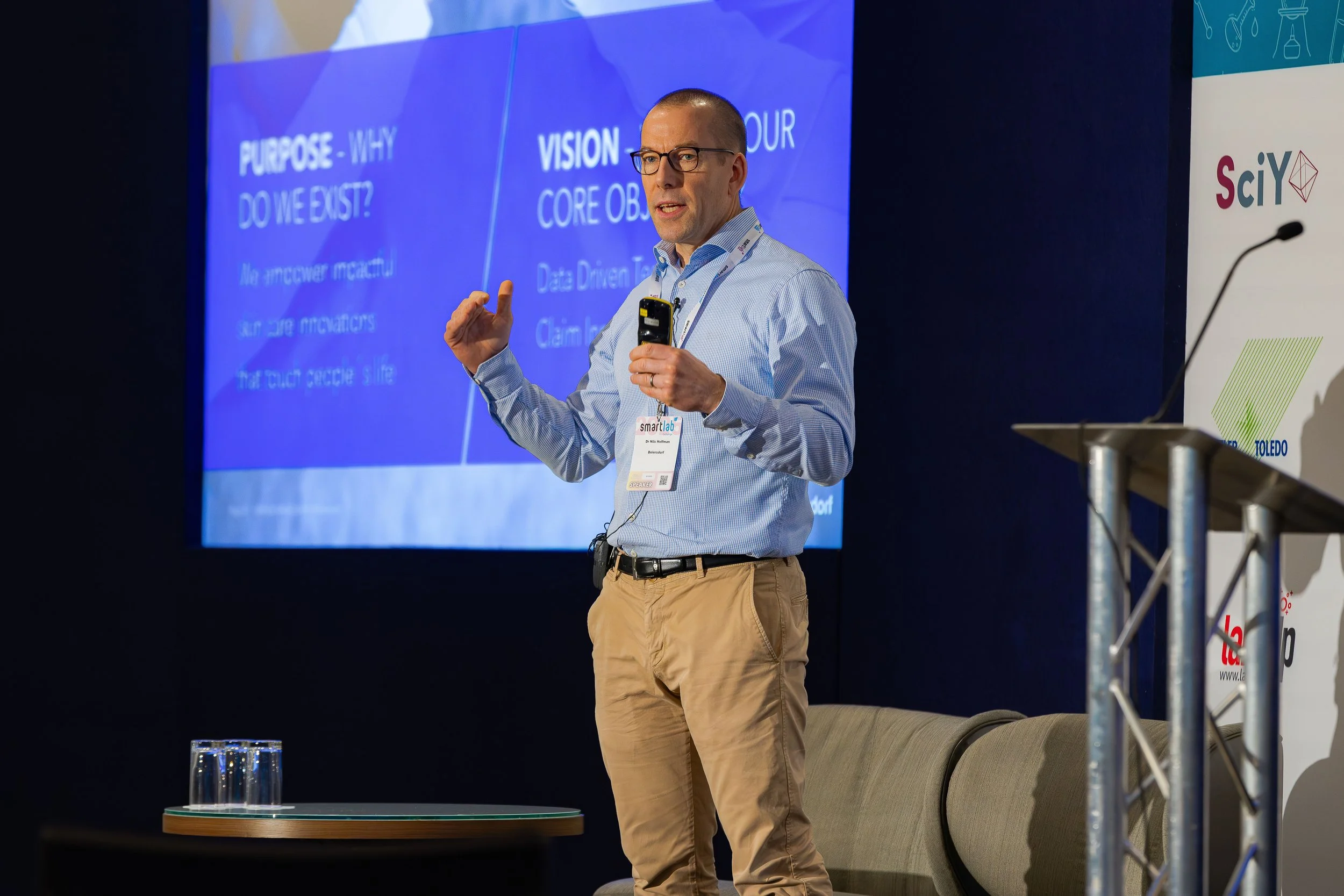 A man with glasses and a blue dress shirt giving a presentation on stage. Behind him is a blue screen with text about purpose, vision, and core objectives. He holds a microphone and points with his index finger. The scene is at a conference or semina