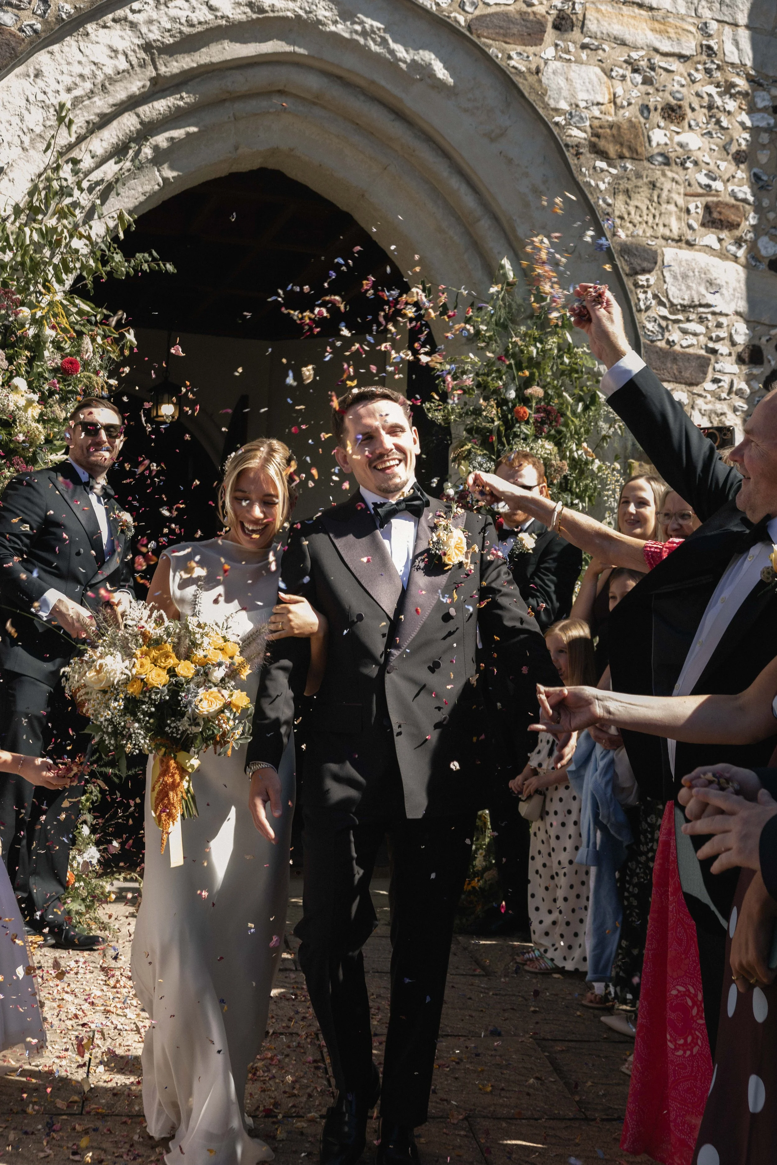 Wedding celebration with a bride in a white dress and a groom in a tuxedo, surrounded by guests throwing flower petals outside a stone building with an arched entrance.