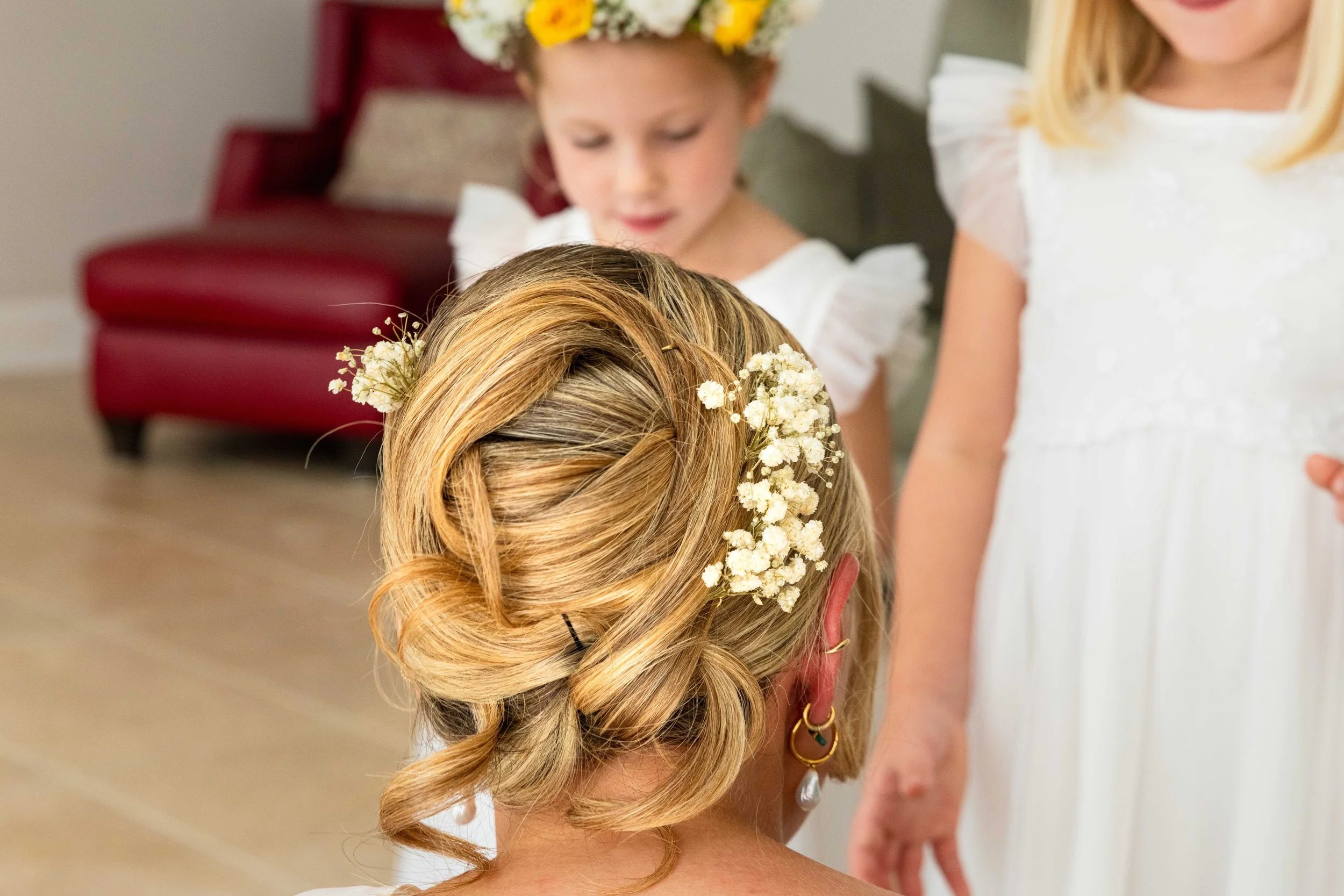 Close-up of a woman's styled hair with blonde curls decorated with small white flowers, indicating a special occasion like a wedding or celebration.