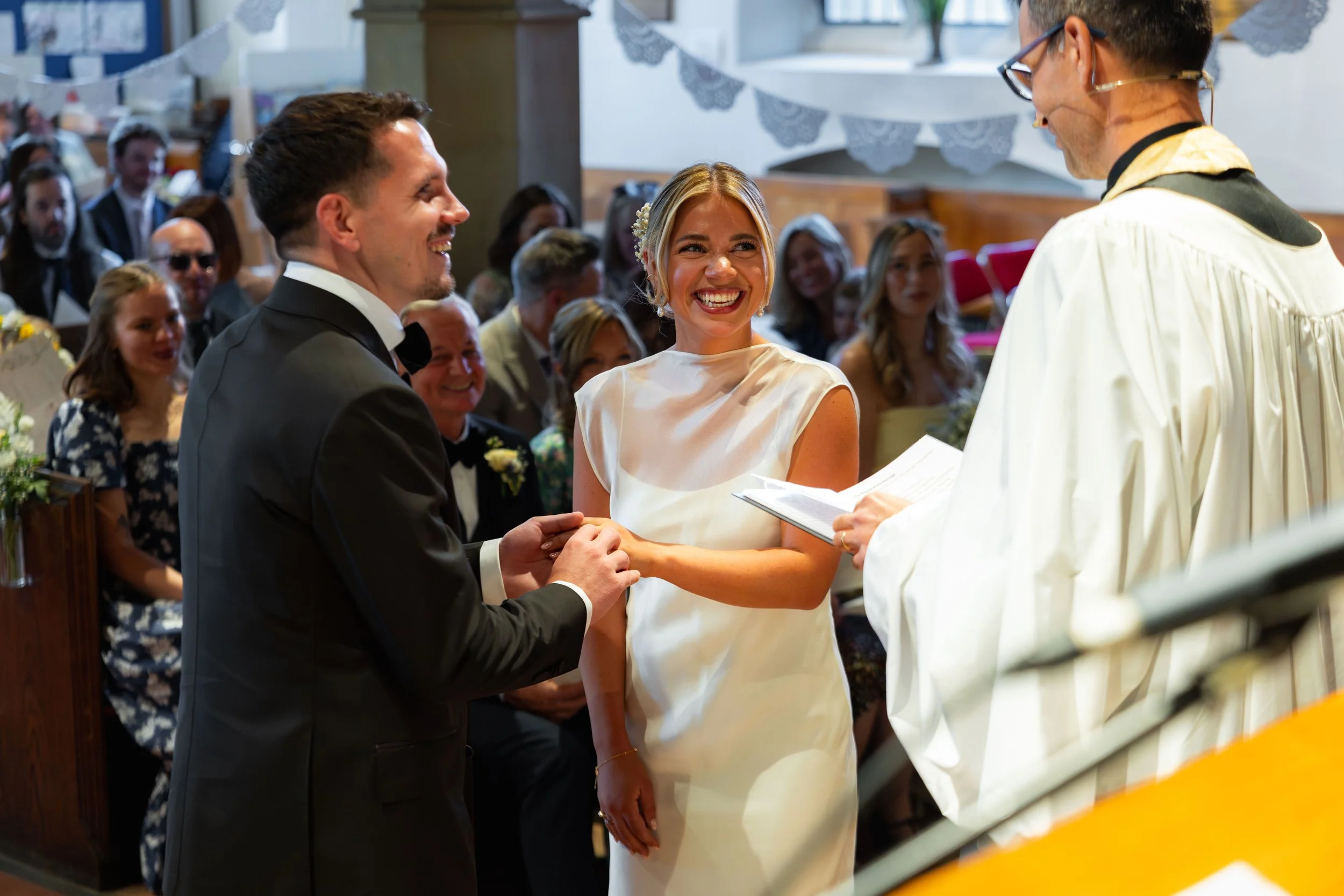 A bride and groom exchanging vows at their wedding ceremony in a church, with an officiant reading from a book and guests watching in the background.