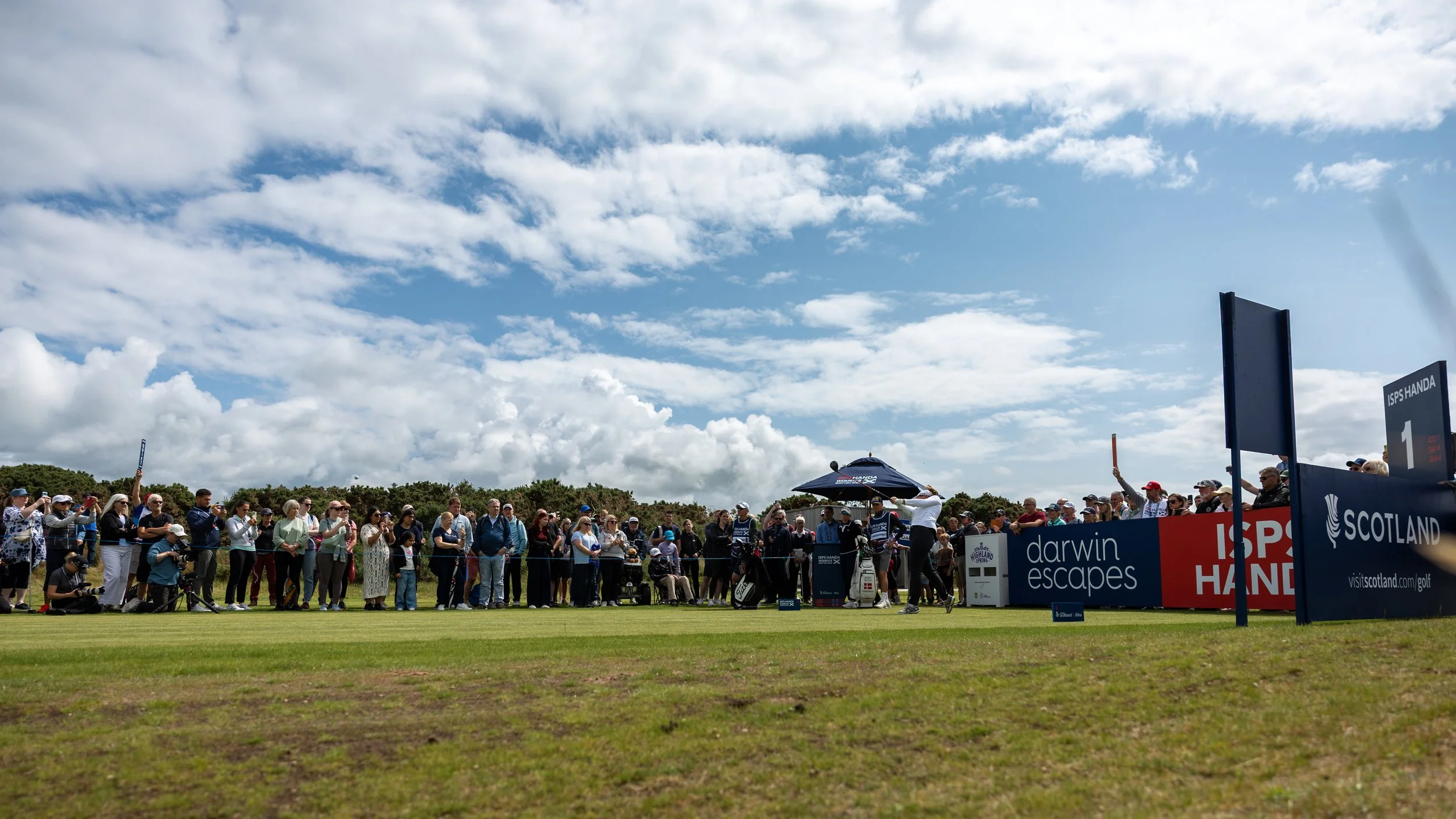 Crowd of spectators at a golf tournament, standing behind a barrier under partly cloudy sky, with sponsor banners including 'Scotland' and 'Darwin Escapes', observing a golfer preparing for a shot.