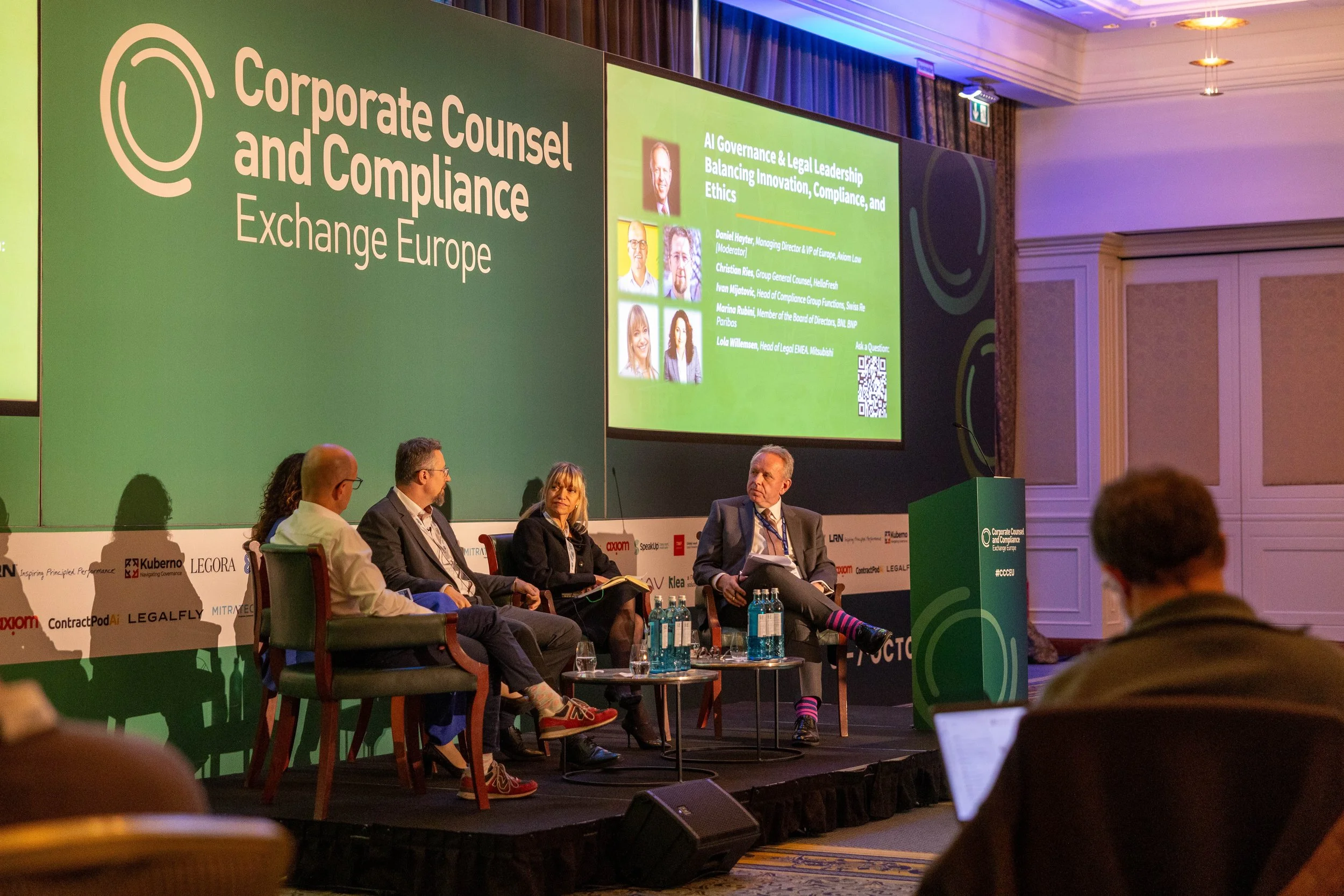 Panel discussion at a conference on corporate counsel and compliance exchange Europe, featuring five panelists seated on stage, with a large green backdrop displaying event details and speaker photos.