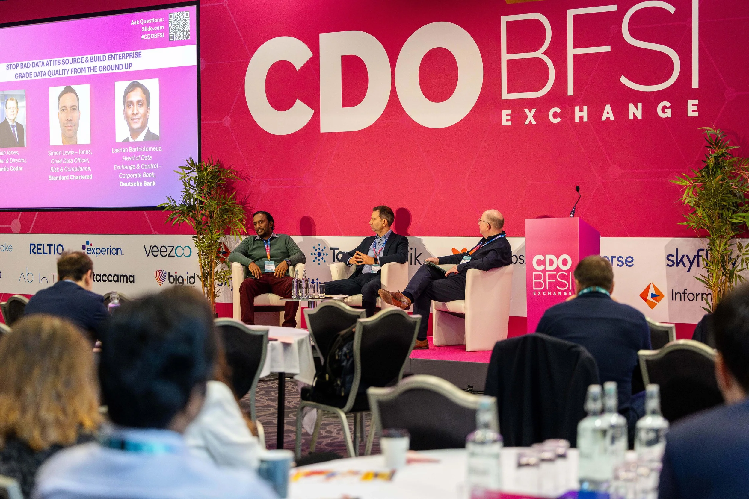A panel discussion at the CDO BFSI Exchange conference. Three men seated on stage in front of a pink backdrop with the event logo, engaging with the audience. The audience members are seated, some taking notes. The stage features a large screen displ
