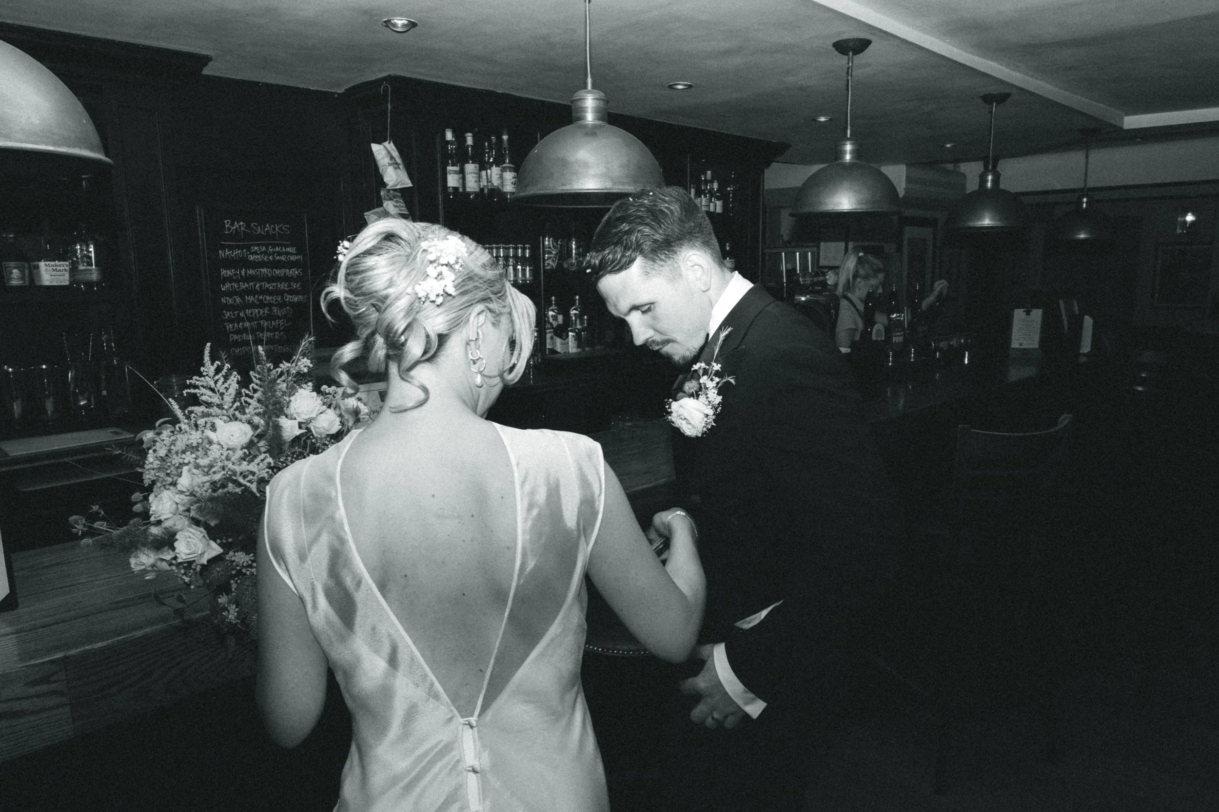 Black and white photo of a bride and groom at a bar. The bride's back is visible, and she is wearing a satin dress with a deep back and floral hair accessories. The groom is kneeling and holding her hand, dressed in a tuxedo with a boutonniere. There