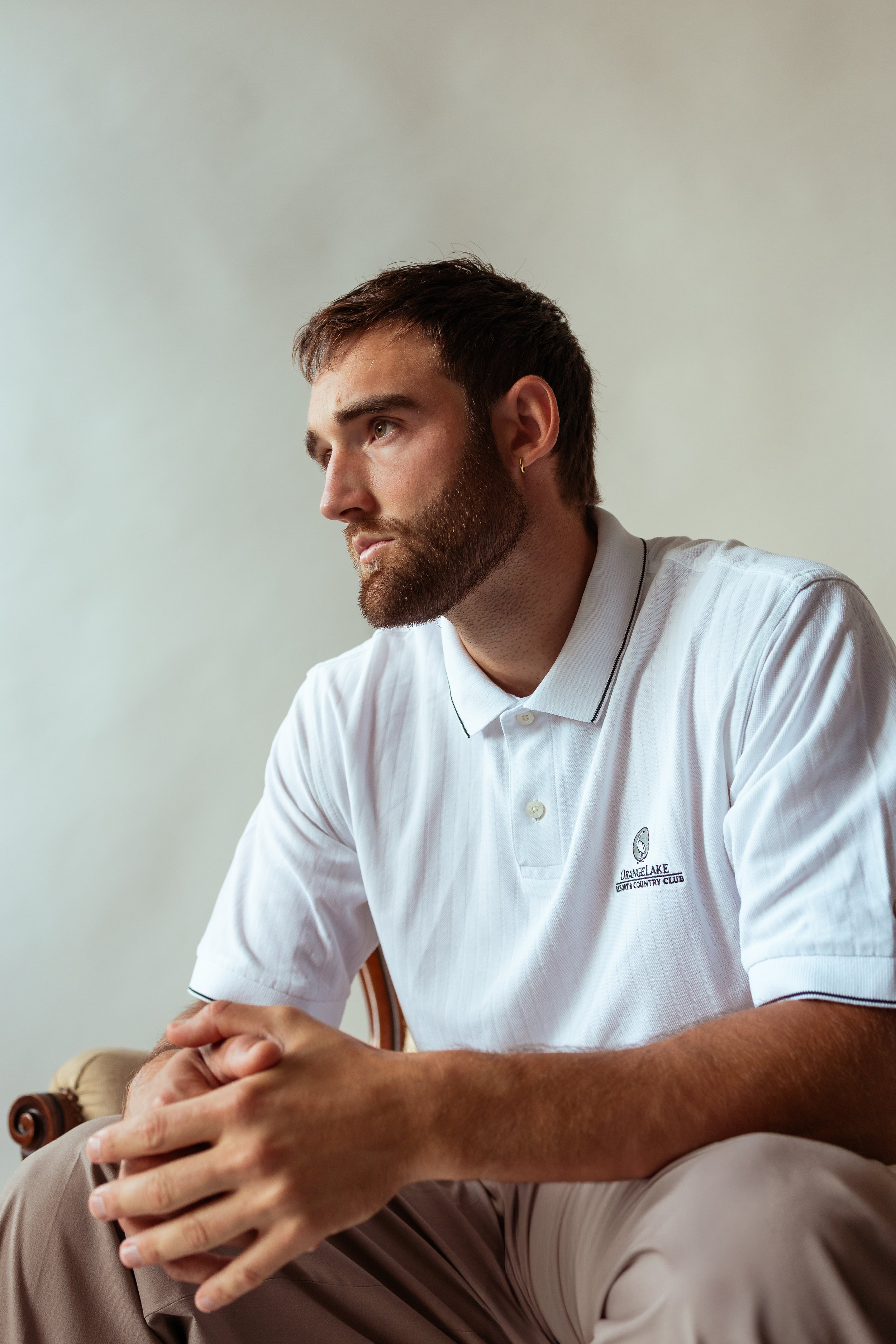 A young man with beard and short hair wearing a white polo shirt with an embroidered logo sitting with his hands clasped and looking to the side against a neutral background.