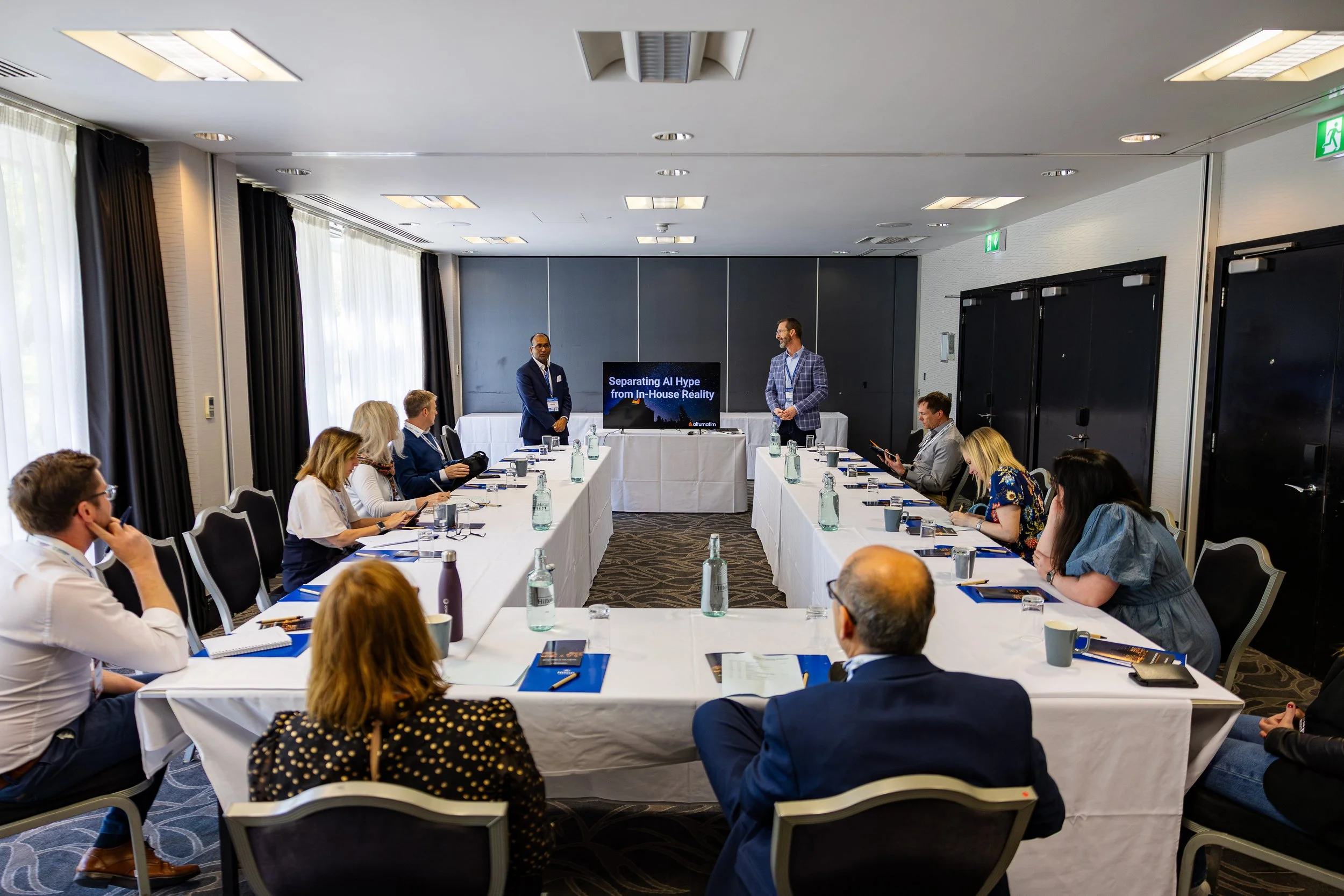A conference room with people seated around U-shaped tables, two men standing at the front giving a presentation on an AI topic, with a screen displaying the title "Separating AI Hype from In-House Reality."