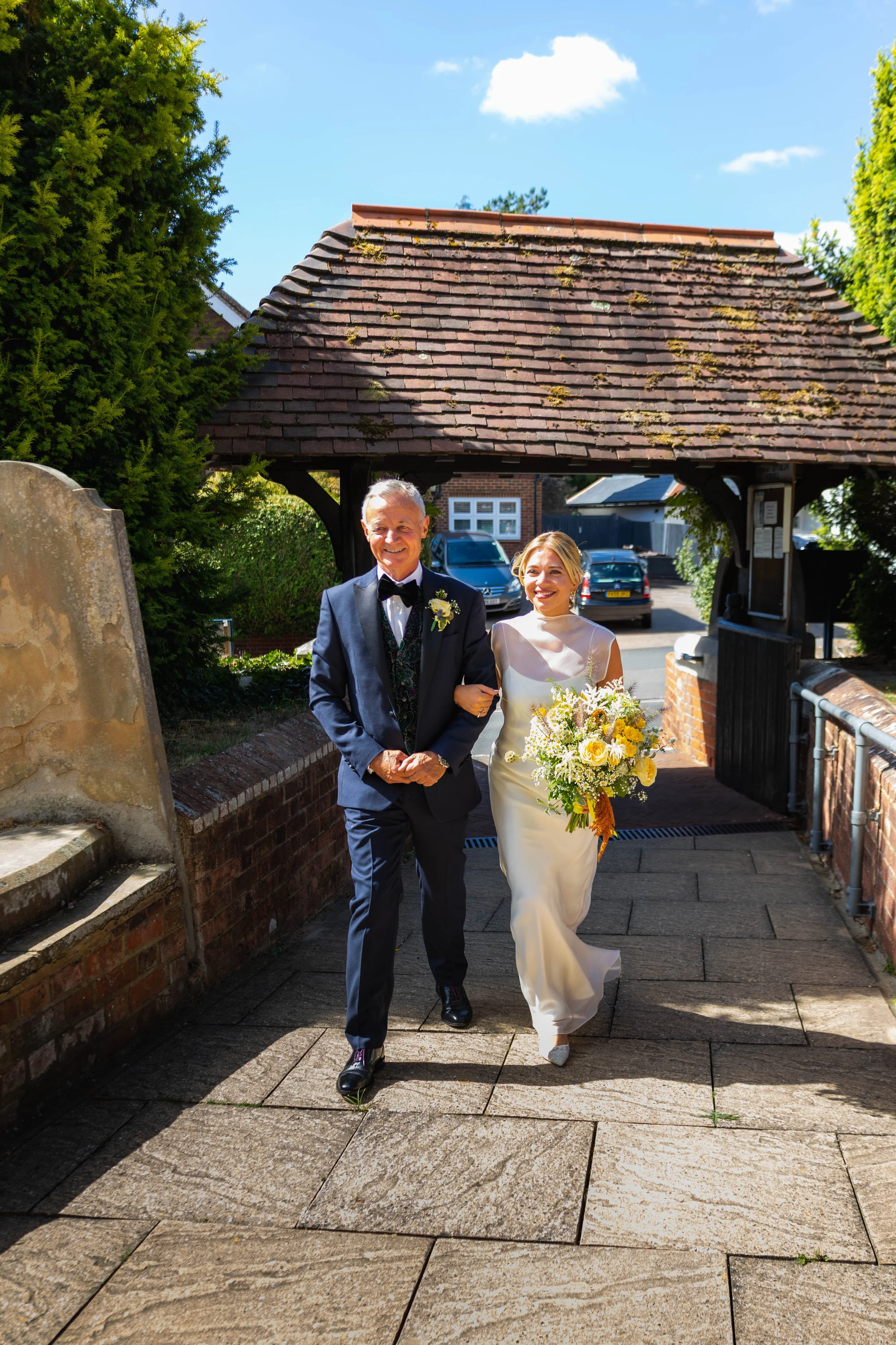 A bride in a wedding dress holding a bouquet walking arm-in-arm with an older man in a tuxedo, outside under a covered walkway with a tiled roof, greenery, and a few cars in the background.