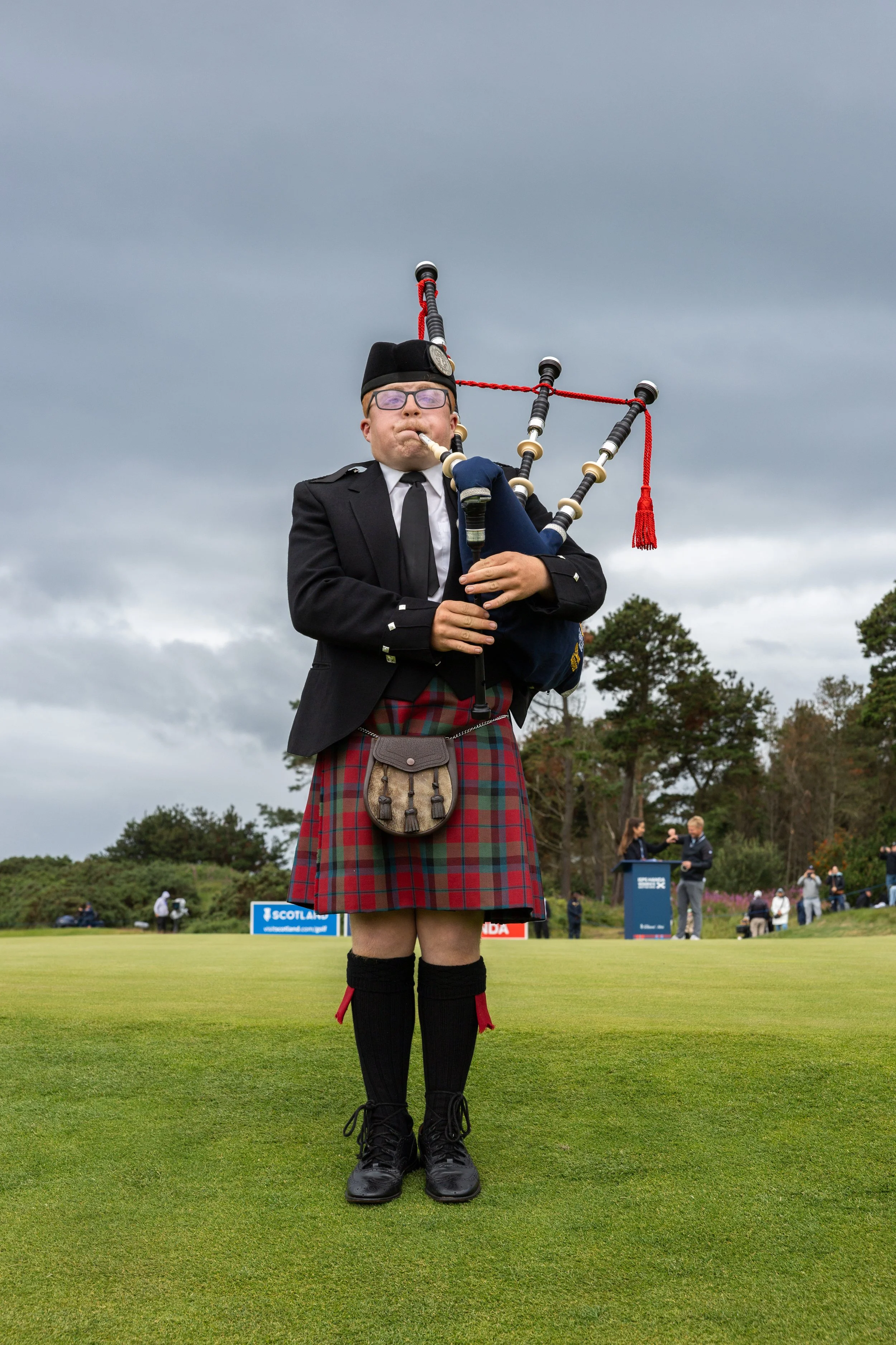 A person dressed in traditional Scottish attire playing bagpipes on a golf course, with spectators and trees in the background under cloudy skies.