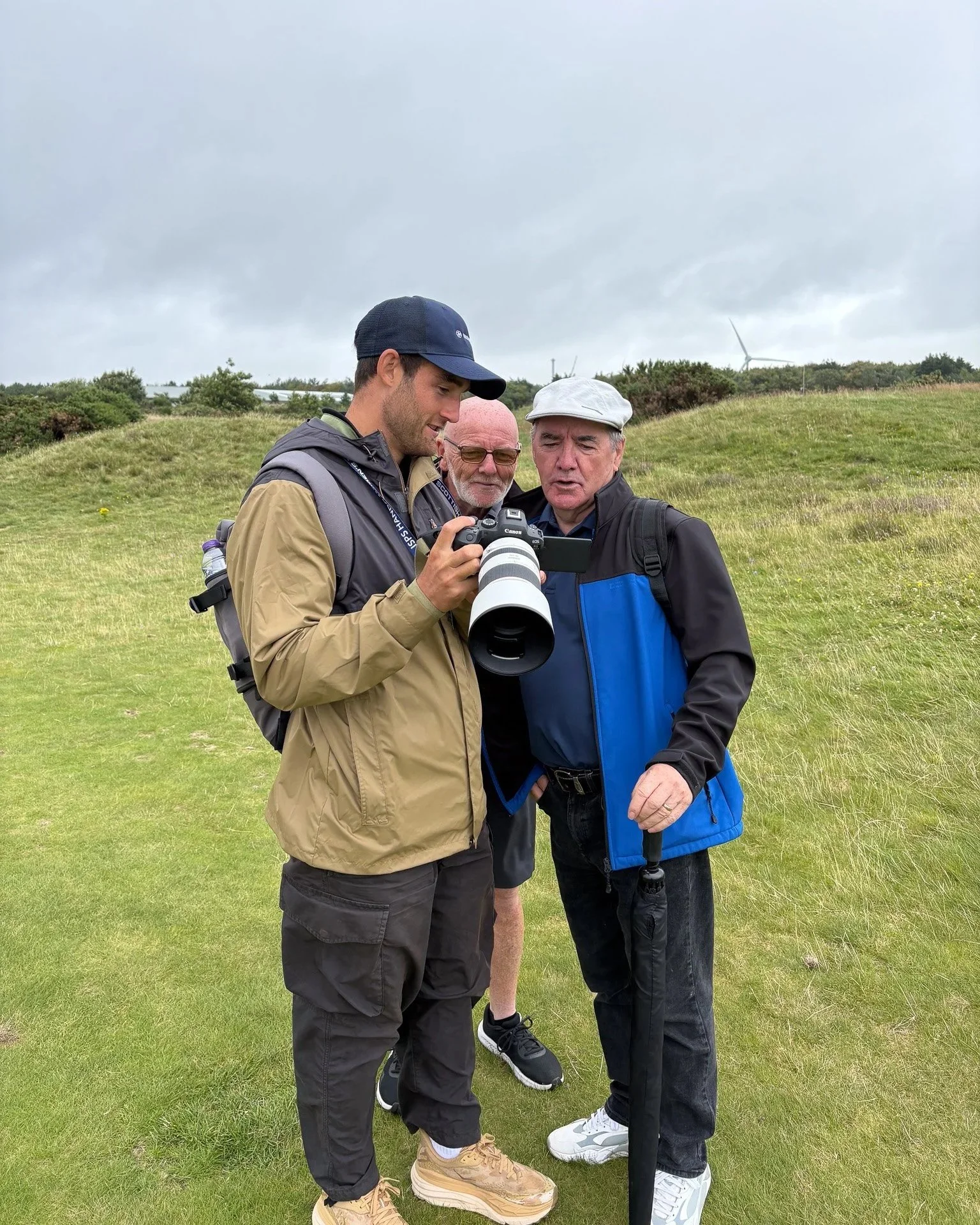 Three men standing outdoors on a grassy hill, looking at a camera held by one of them. The background shows cloudy skies and wind turbines.