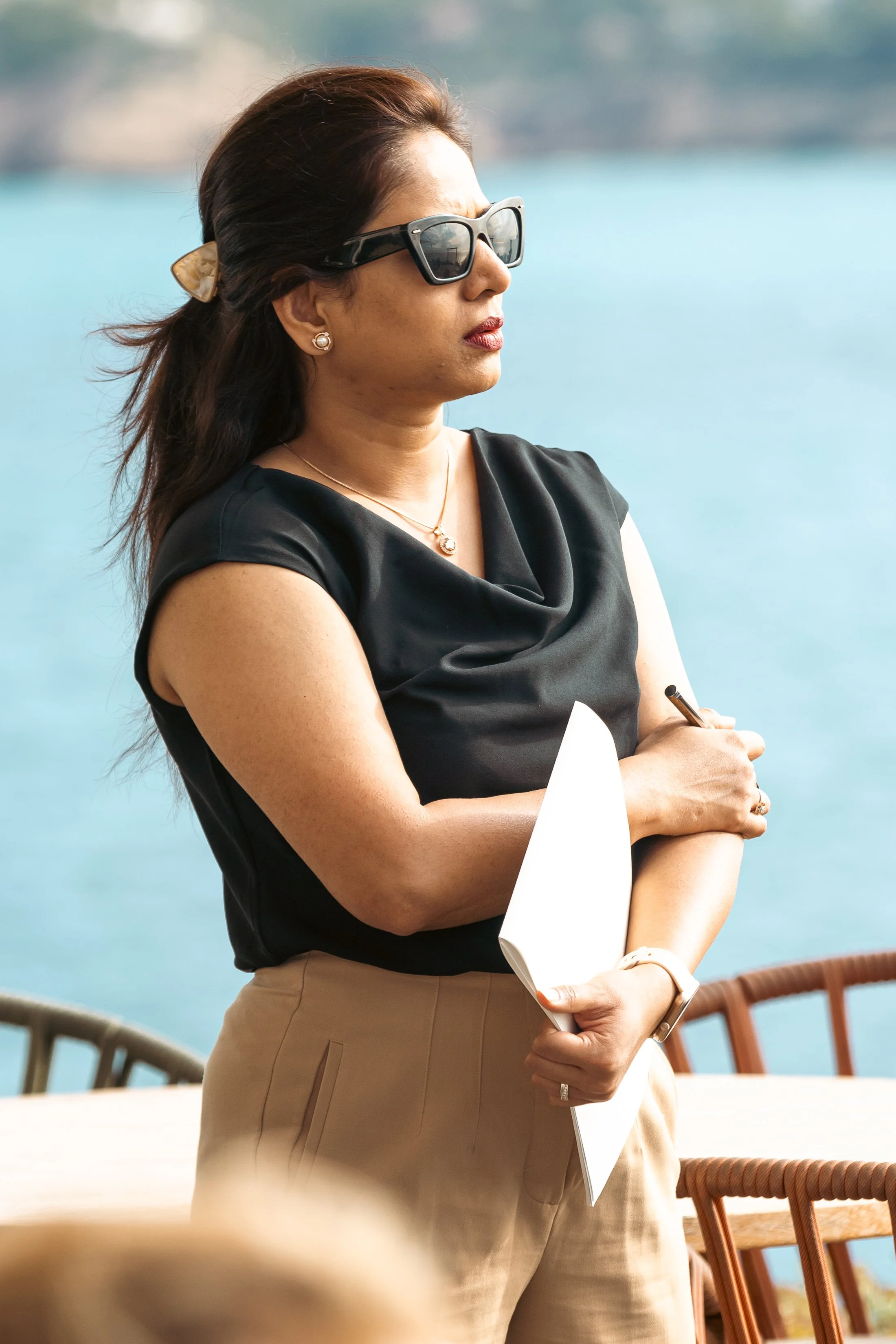 A woman with brown hair wearing black sunglasses, pearl earrings, a gold necklace, and a black top standing outdoors near a body of water, with her arms crossed holding a paper and pen.
