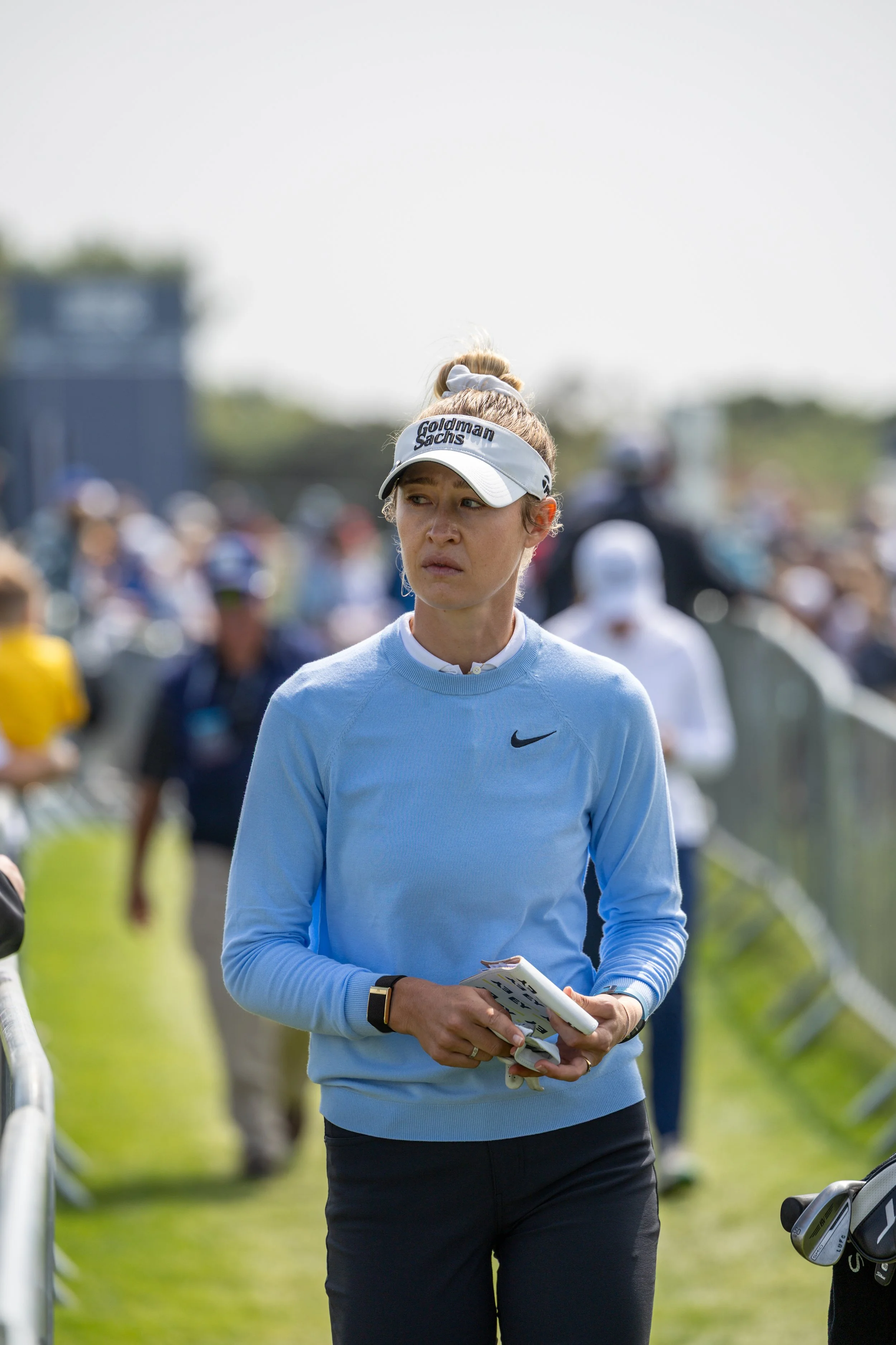A female golfer wearing a light blue Nike sweatshirt and a white visor with 'Goldman Sachs' on it, standing outdoors at a golf event, holding a pamphlet, with blurred spectators and golf equipment in the background.