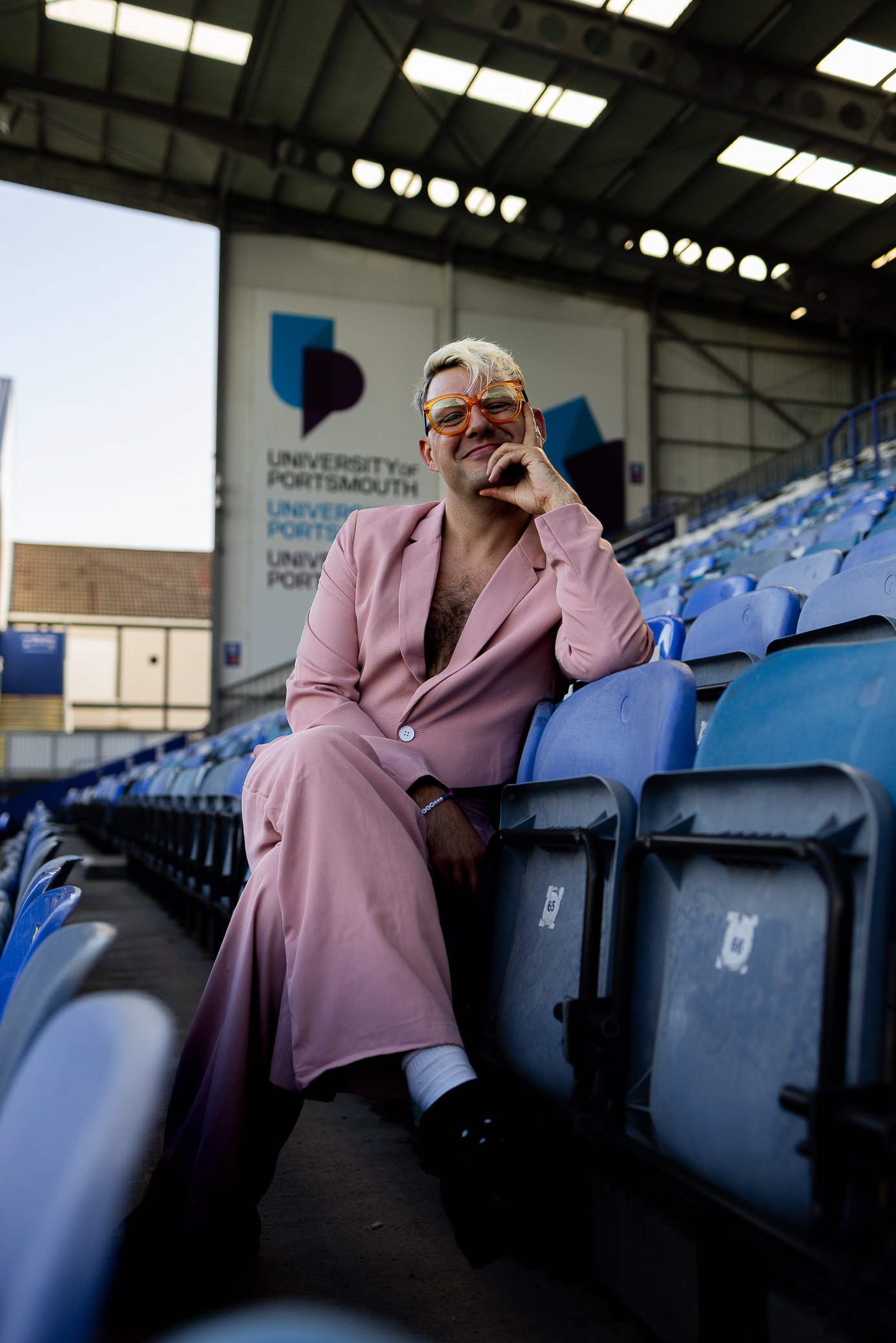 A person with short blonde hair and orange glasses sitting in empty blue stadium seats under a roof with large windows, wearing a pink suit with an unbuttoned shirt, smiling and resting their chin on their hand, with a banner in the background that r