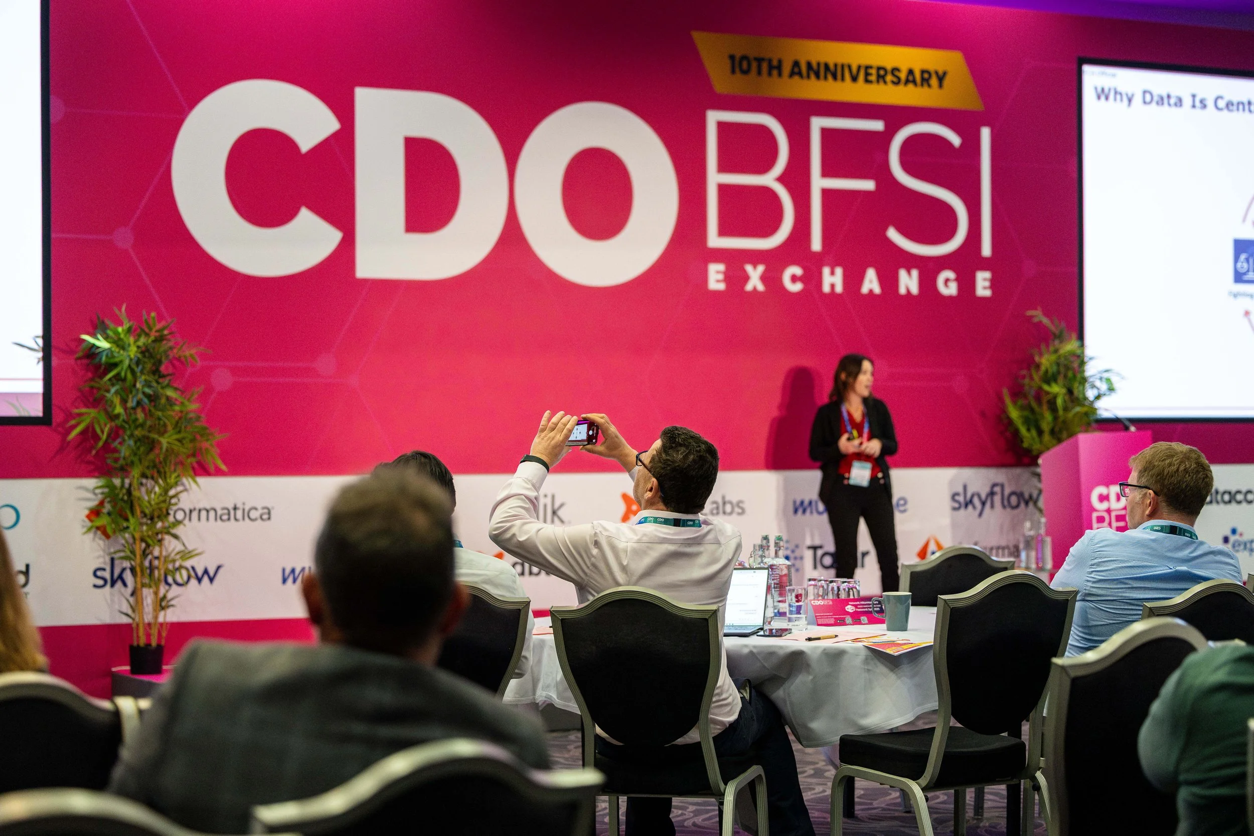 Conference room with a large pink banner reading 'CDO BEs Exchange 10th Anniversary'. Several attendees are seated at tables, some taking photos of a woman presenting near a screen, with potted plants on either side of the stage.