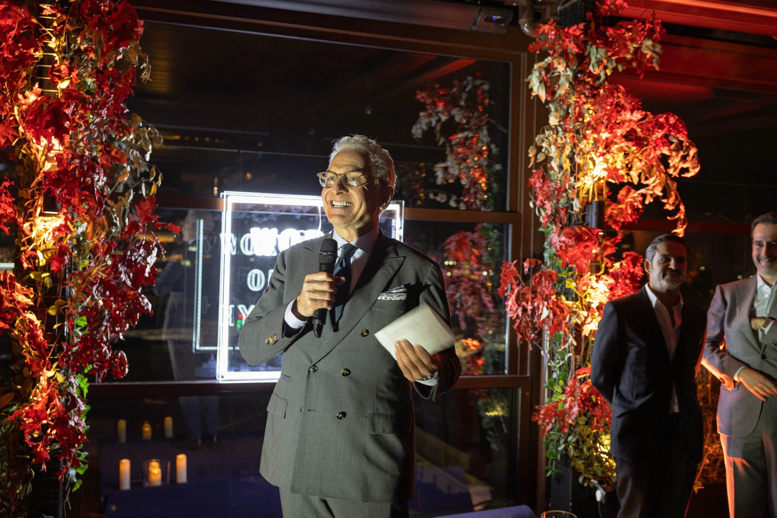 A man in a gray suit and glasses holding a microphone and paper, standing in front of a decorated background with red leaves and flowers during an event at night.