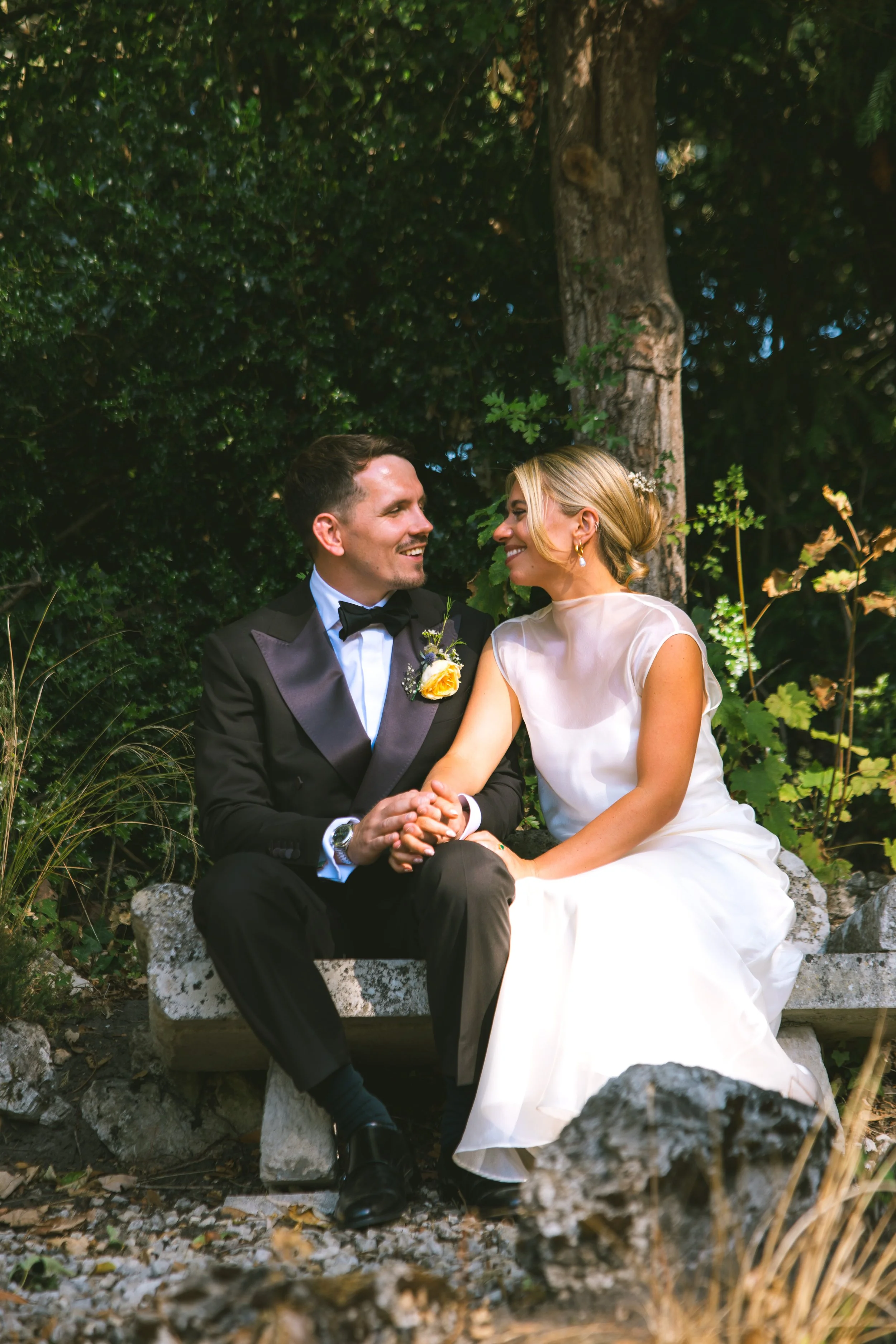 A groom and bride sitting on a stone bench outdoors, holding hands and smiling at each other surrounded by green foliage.