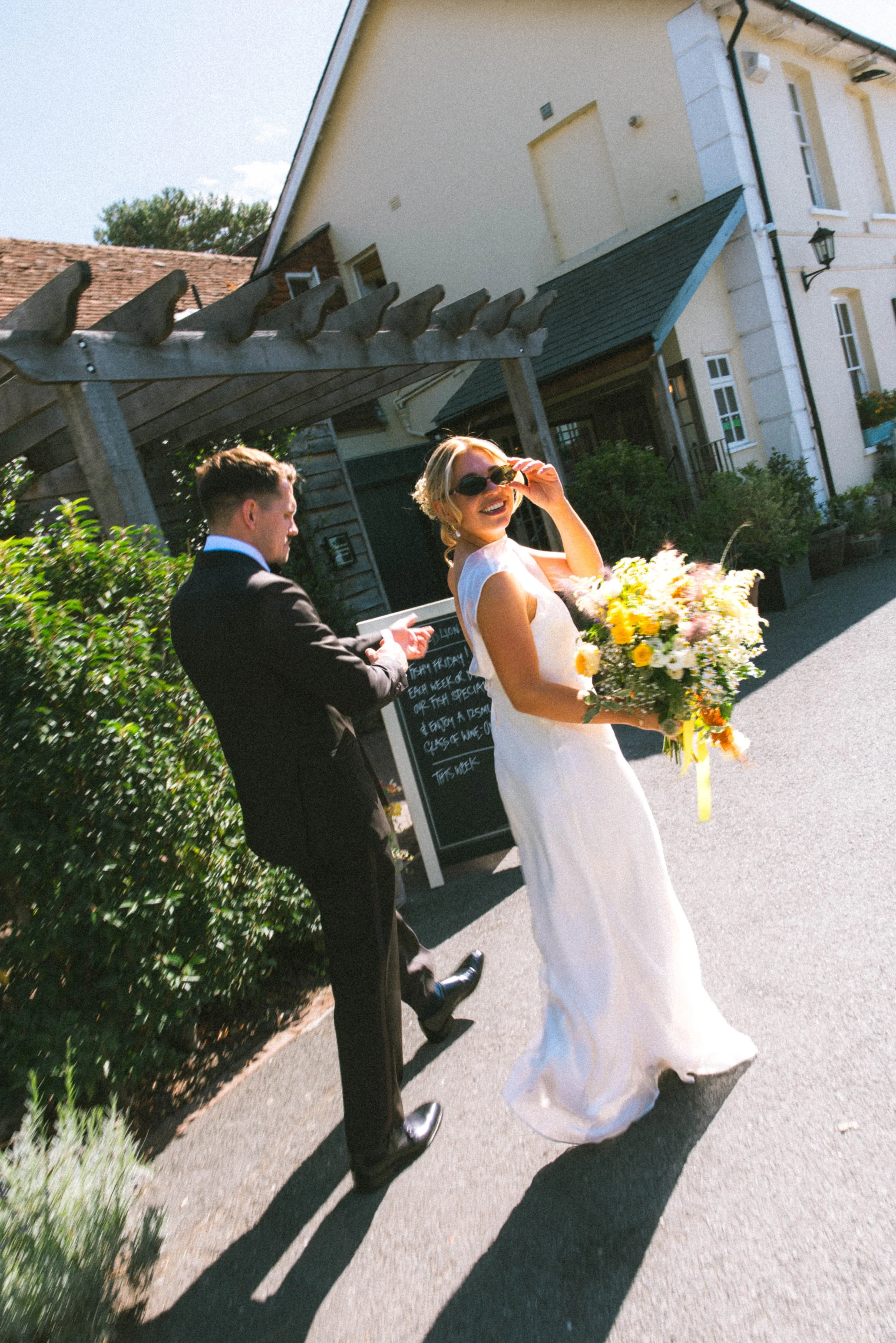 A woman in a white wedding dress holding a bouquet of flowers, smiling and wearing sunglasses, walking outdoors. A man in a black suit is behind her, also walking outdoors. The background shows a white building with black lanterns and greenery.