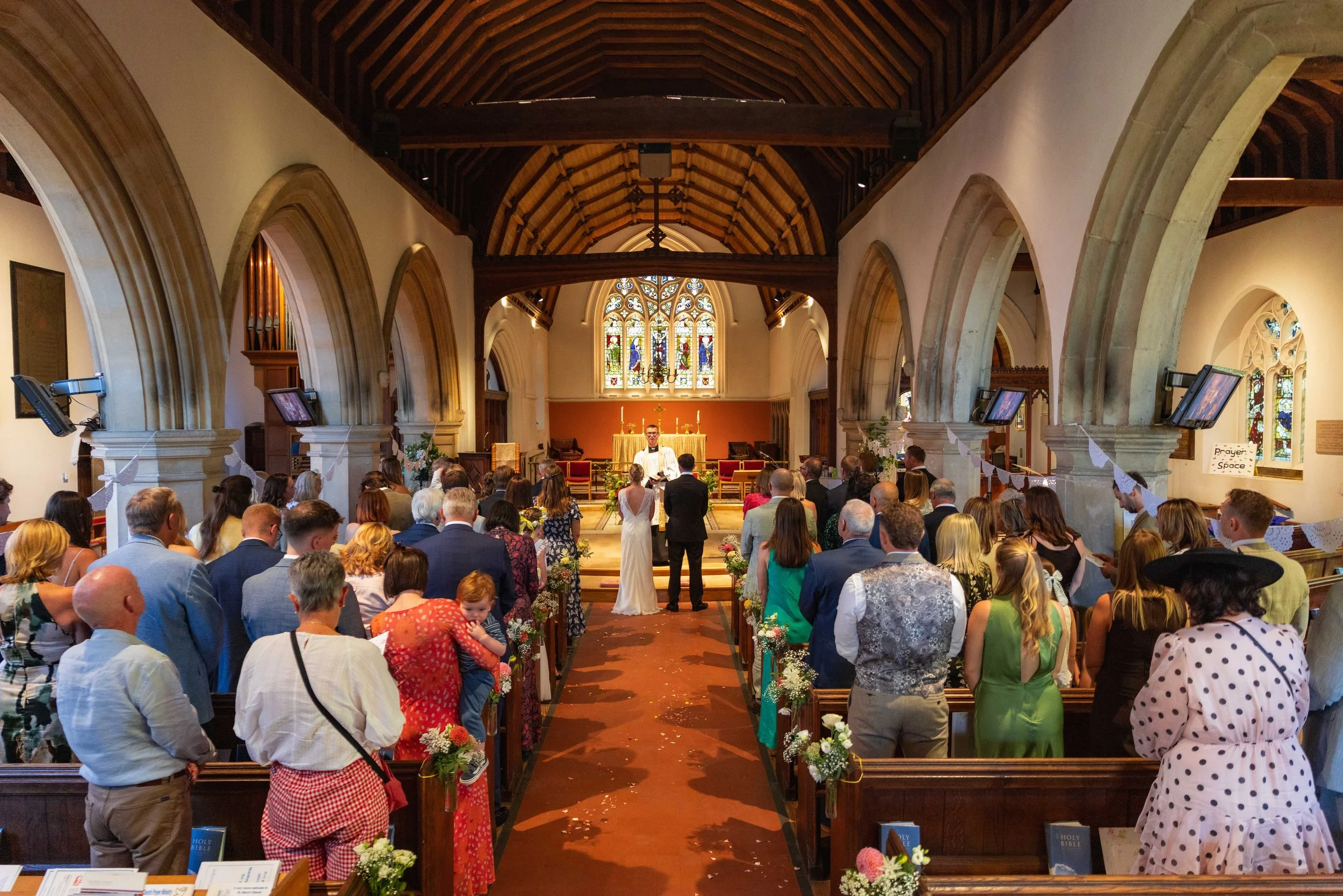 A wedding ceremony inside a church with many guests, a bride and groom at the altar, and a minister officiating. The church has stained glass windows, wooden ceiling beams, and pews filled with attendees.