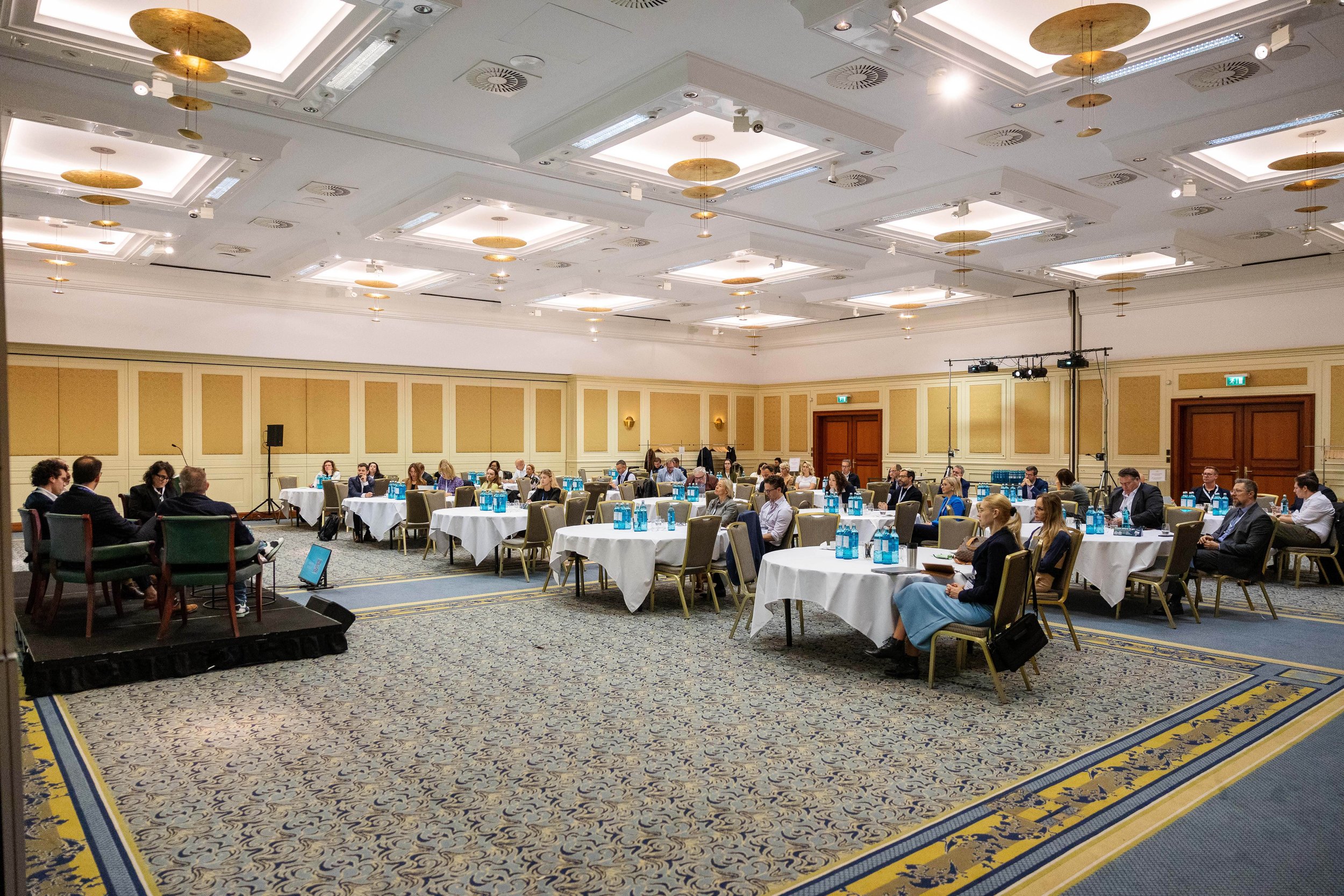 Large conference room filled with people seated at round tables, each with blue water bottles and notepads. The room features a high ceiling with decorative gold circular fixtures, beige walls with paneling, and a patterned carpet. There is a speaker