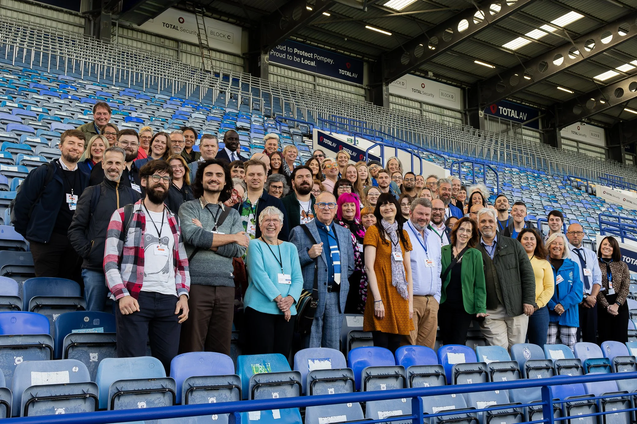 A large group of people posing for a photo inside a stadium with blue seats. The group includes men and women of various ages and ethnicities, all smiling. Some are wearing jackets, and many have conference badges around their necks. The stadium is m
