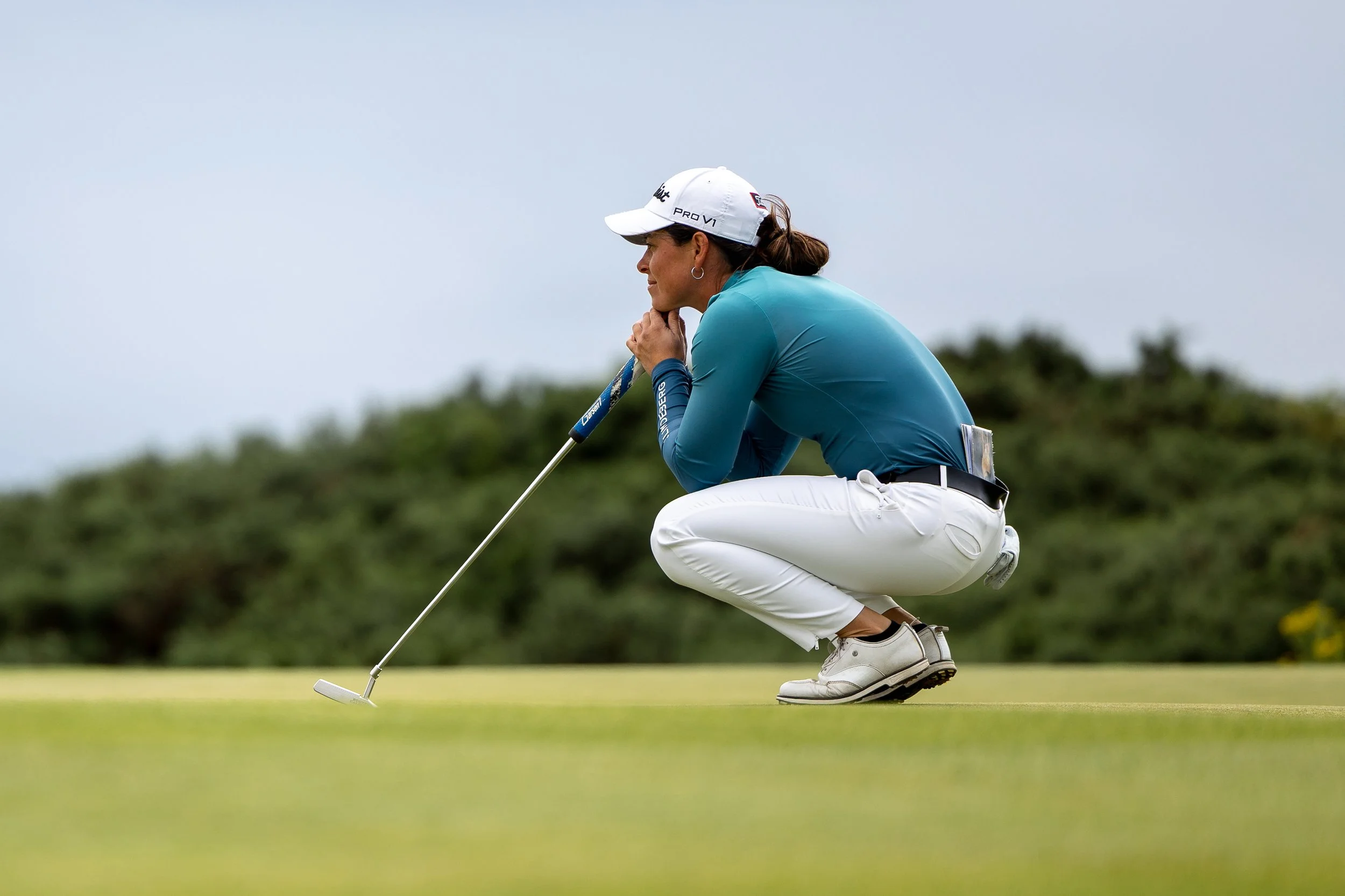 A female golfer is crouching on a golf course, holding a putter in her right hand, wearing a white cap, teal shirt, white pants, and white shoes, with greenery in the background.