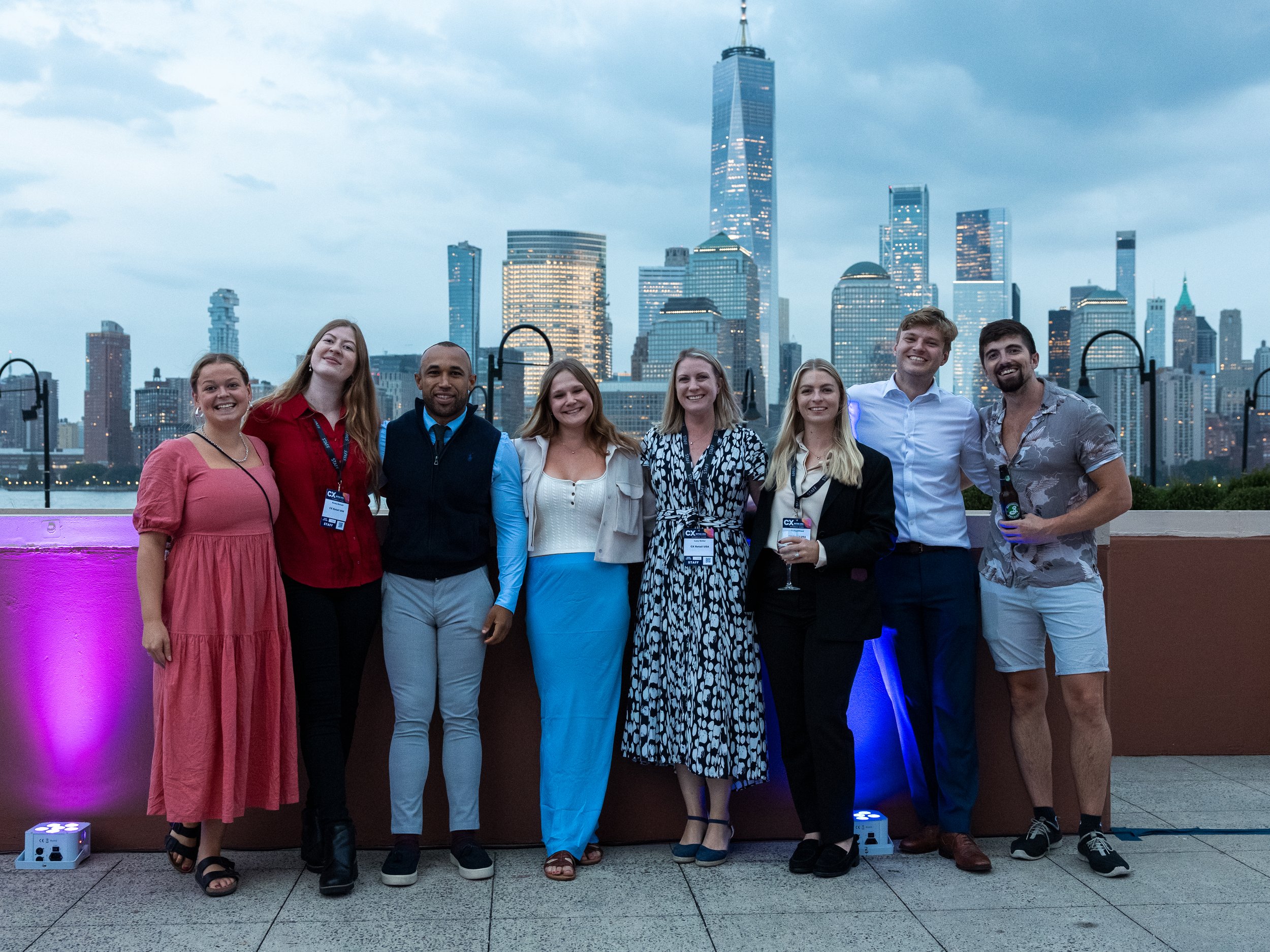 A group of eight diverse young adults standing together on a rooftop with the New York City skyline in the background during evening hours, smiling and posing for a photo.