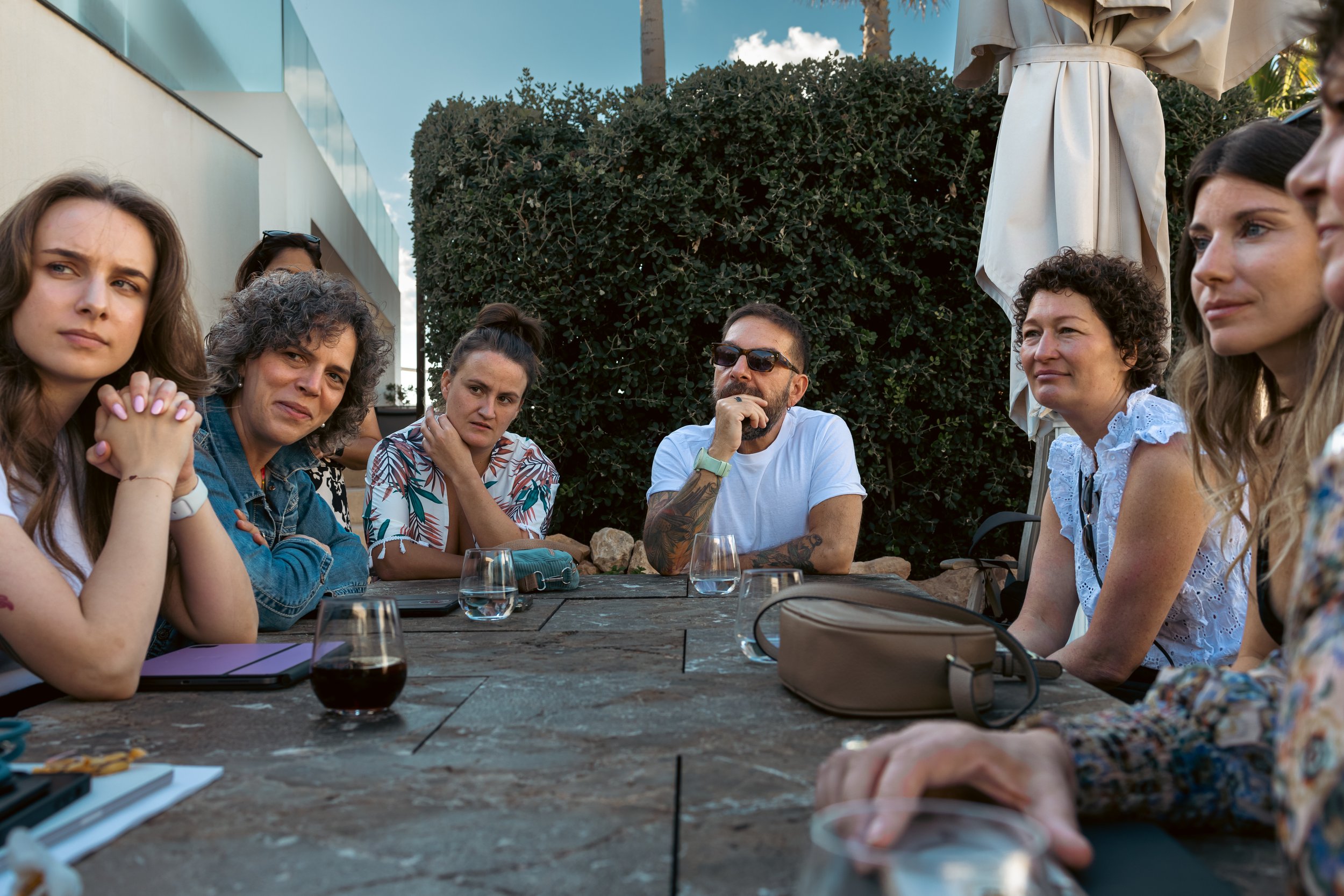 Group of diverse people sitting around a table outdoors, engaged in a serious discussion, with a bush and blue sky in the background.
