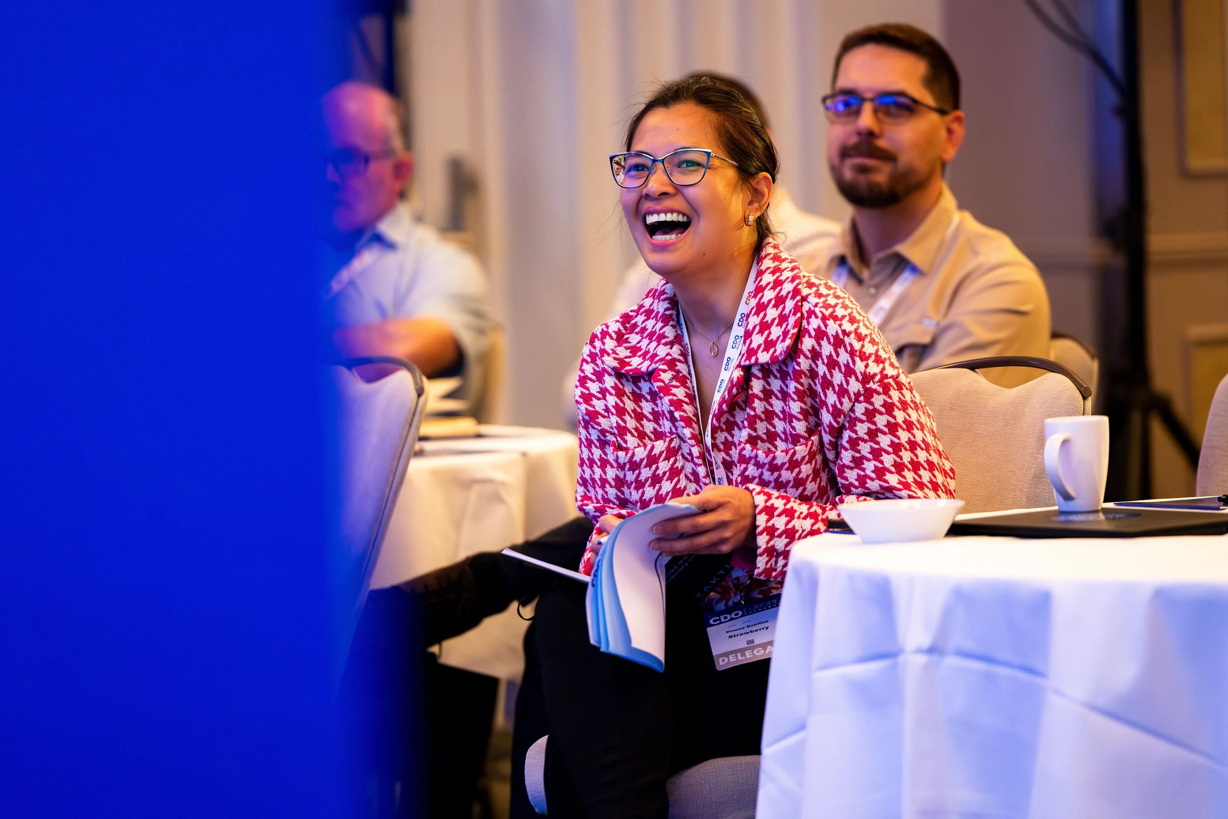 A woman with glasses wearing a red and white patterned jacket, smiling and laughing at a conference or seminar. She is seated at a table with a white tablecloth, with a coffee mug and a bowl in front of her. Other attendees are visible in the backgro