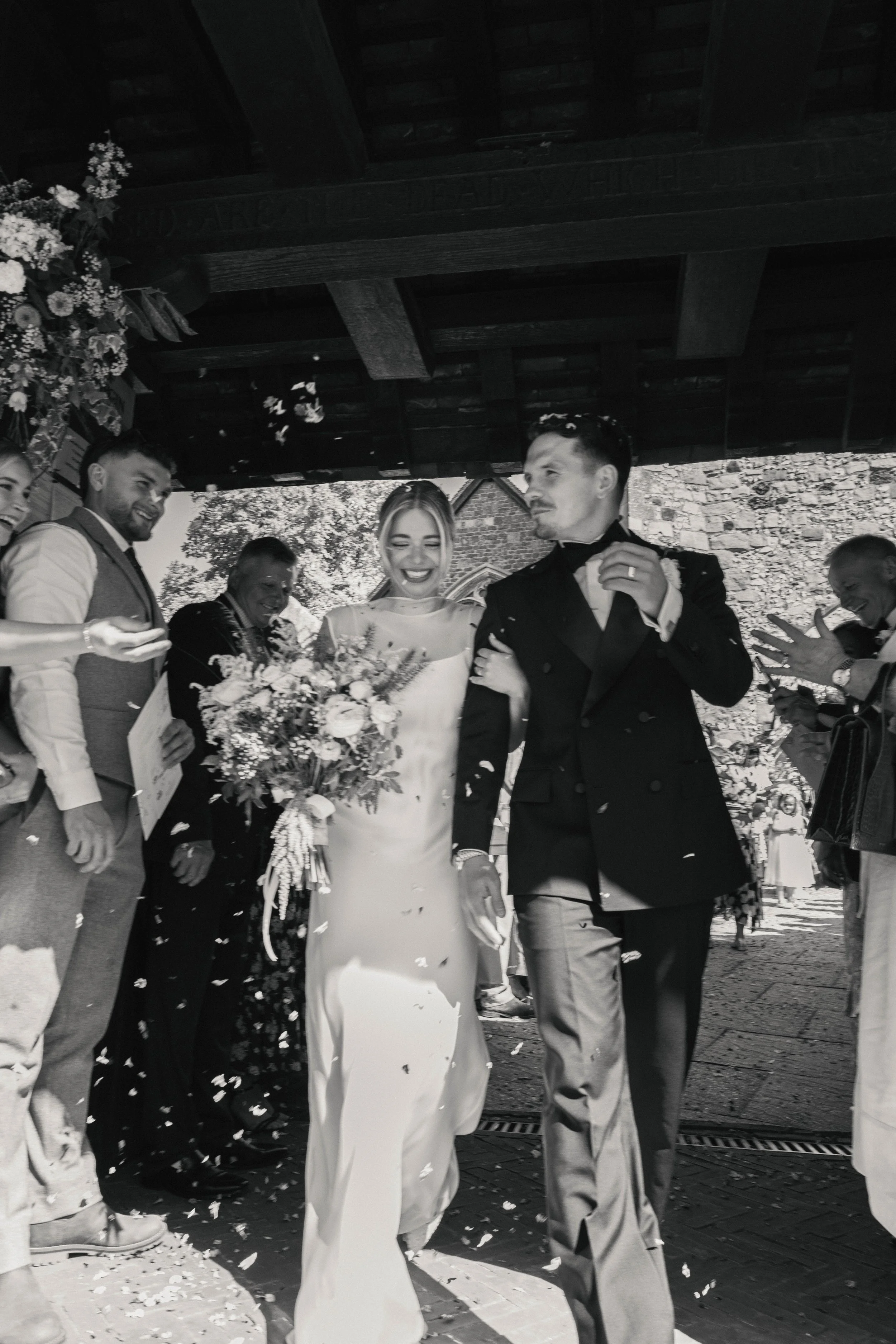 A black and white photo of a wedding celebration showing a bride and groom smiling and holding hands, surrounded by wedding guests. The bride holds a bouquet of flowers, and everyone appears happy, with some guests throwing confetti.