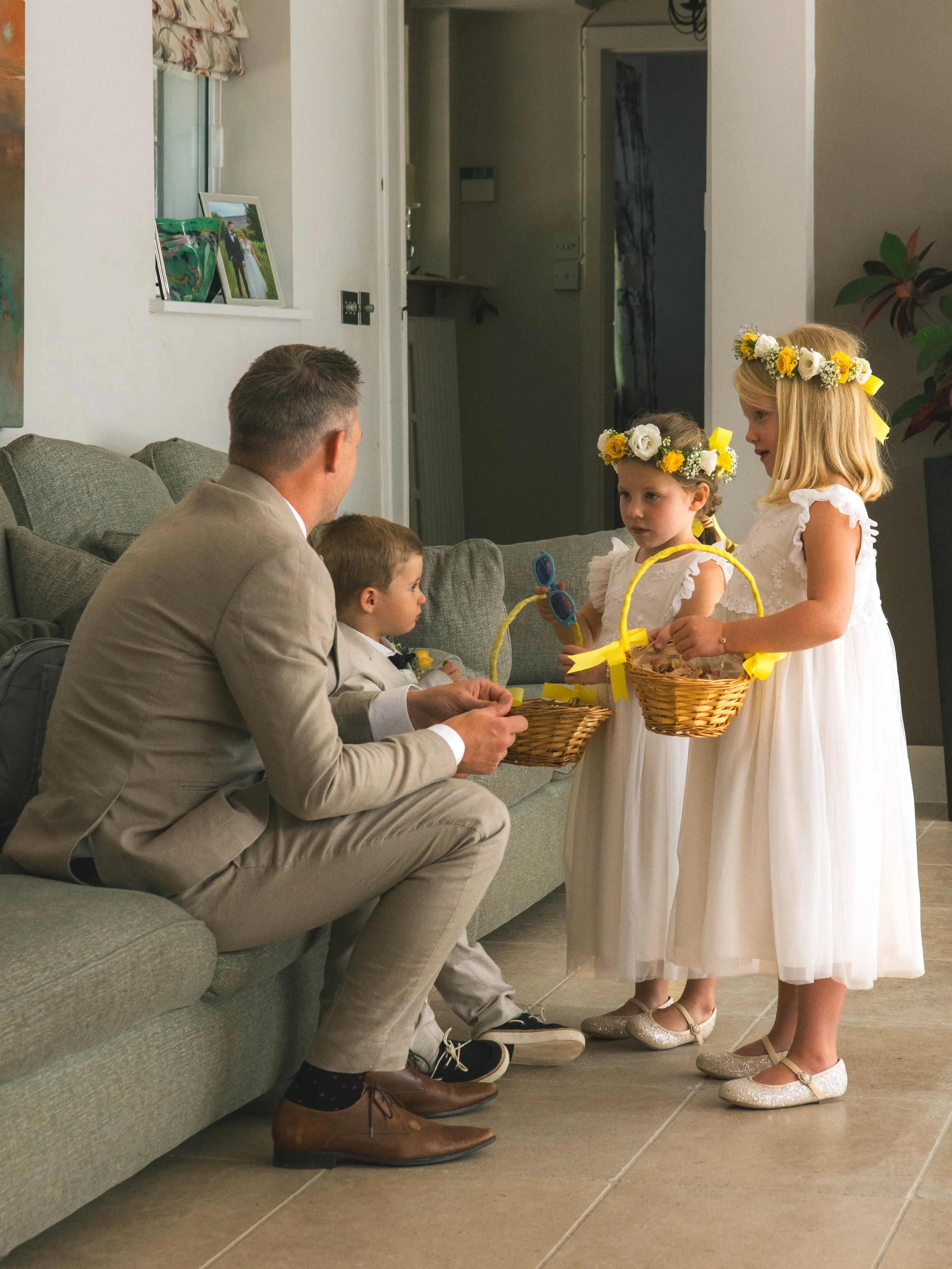 Two young girls and a man are standing near a couch, holding baskets and dressed in white dresses with floral headbands, possibly participating in a wedding ceremony or celebration. The man and one girl are seated, while the other girl stands facing 
