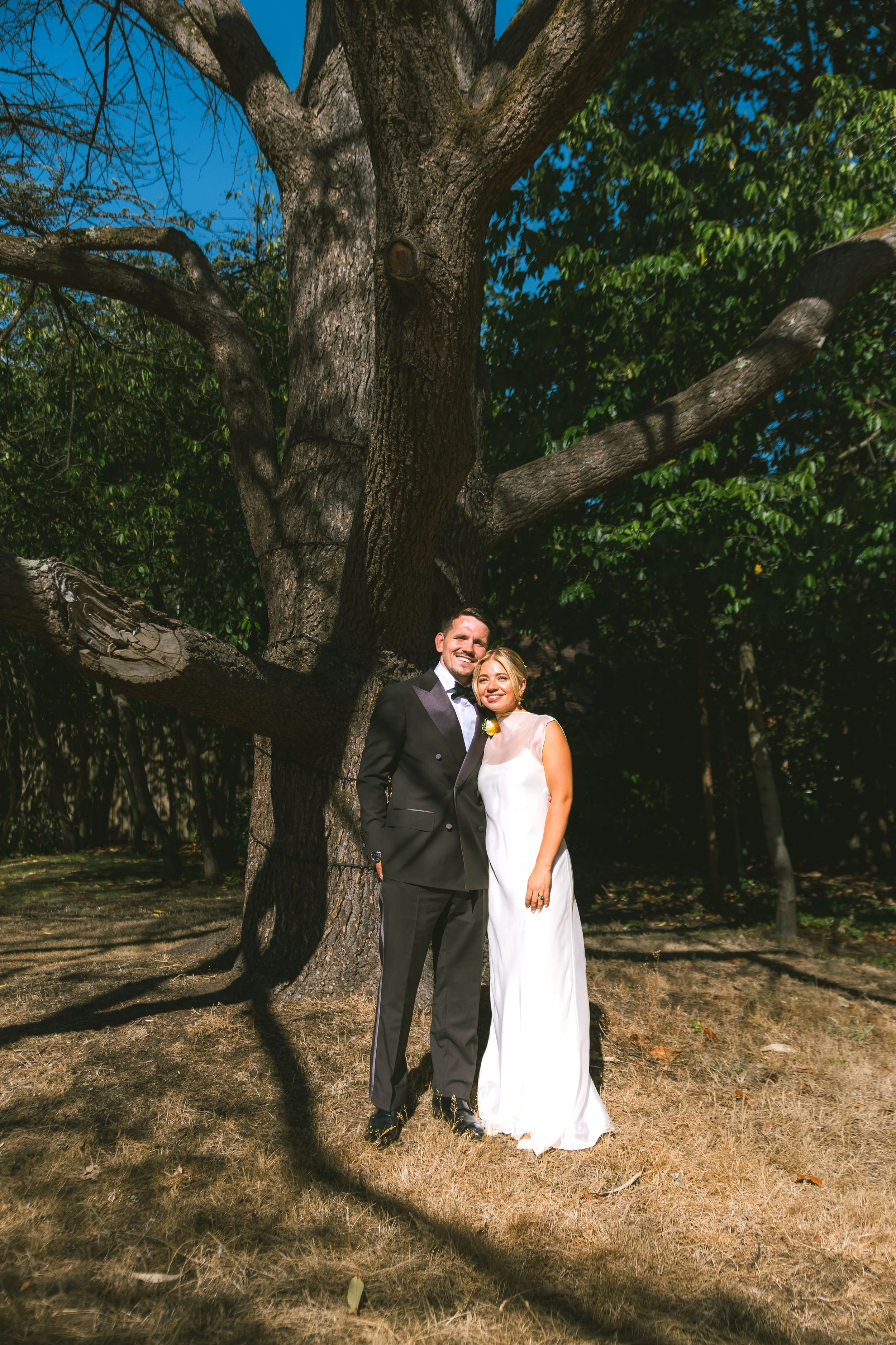 A newlywed couple stands outdoors under a large tree with thick branches and green foliage, smiling at the camera. The groom wears a black tuxedo, and the bride wears a white wedding dress. The scene is taken in a park or garden setting on a sunny da