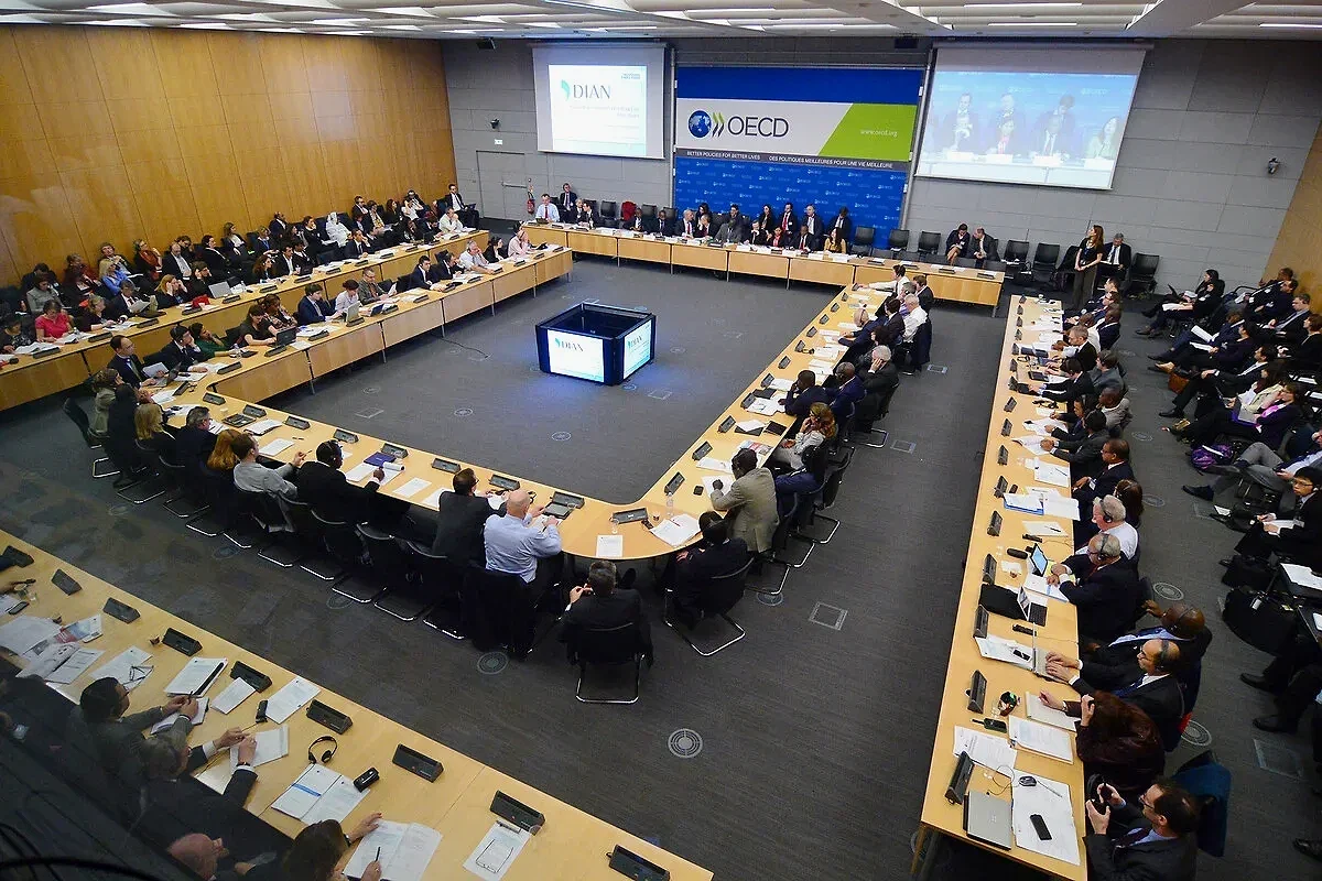 A large conference room with a U-shaped arrangement of tables filled with businesspeople, speakers, and attendees, some wearing headsets, attending an international meeting organized by OECD with a presentation projected on screens at the front.