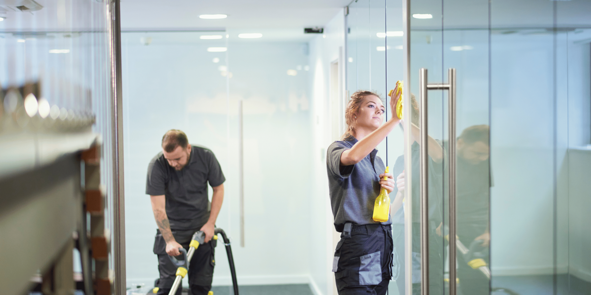 Two cleaning workers, a woman and a man, cleaning a glass wall in an office building. The woman is using a yellow spray bottle and cloth, while the man is vacuuming the carpet.