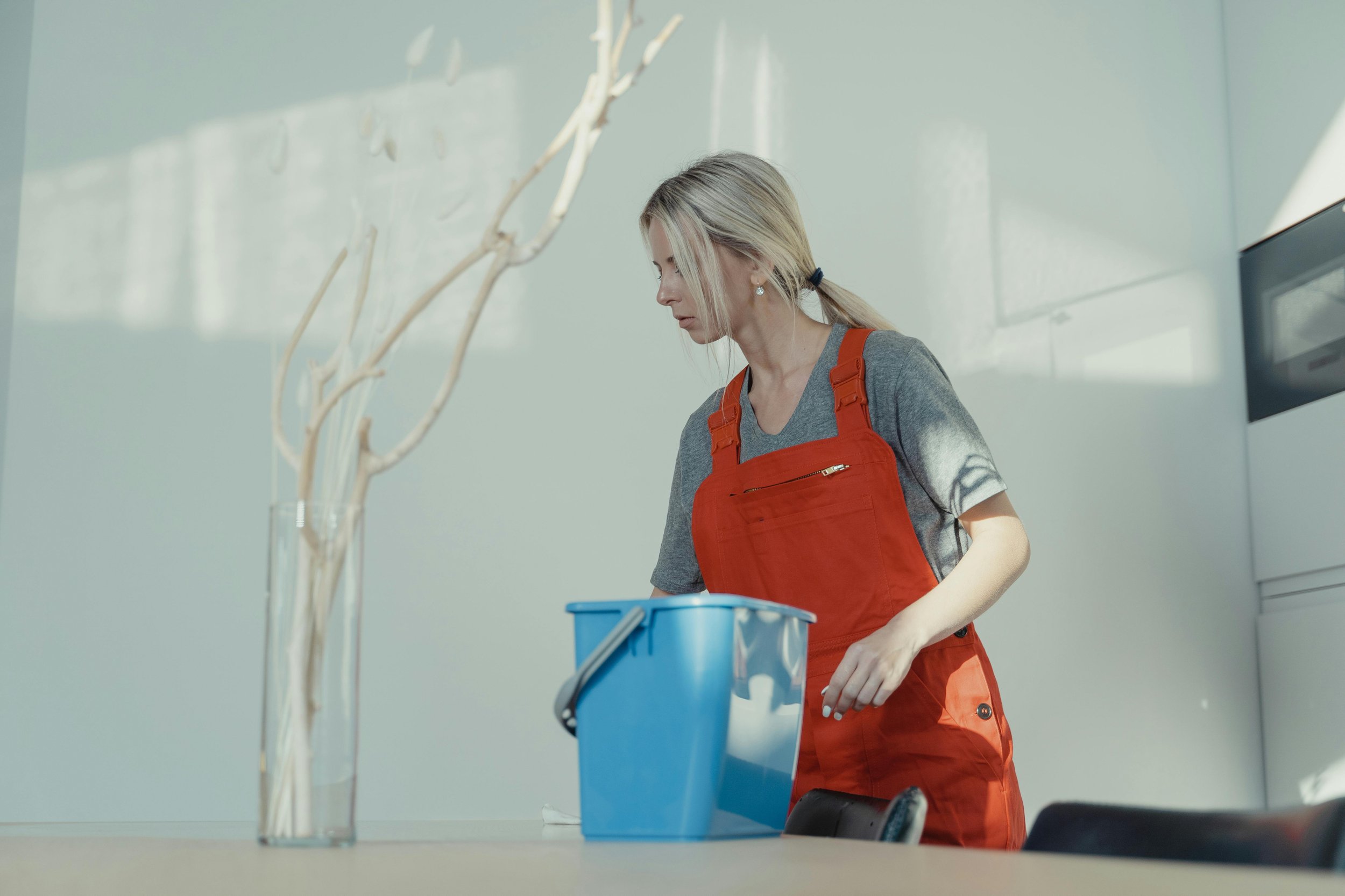 A woman with blonde hair tied back, wearing diamond earrings, a gray t-shirt, and an orange apron, standing outdoors near a white wall and a window, with a blue plastic bucket and a clear vase with decorative branches on a table in front of her.