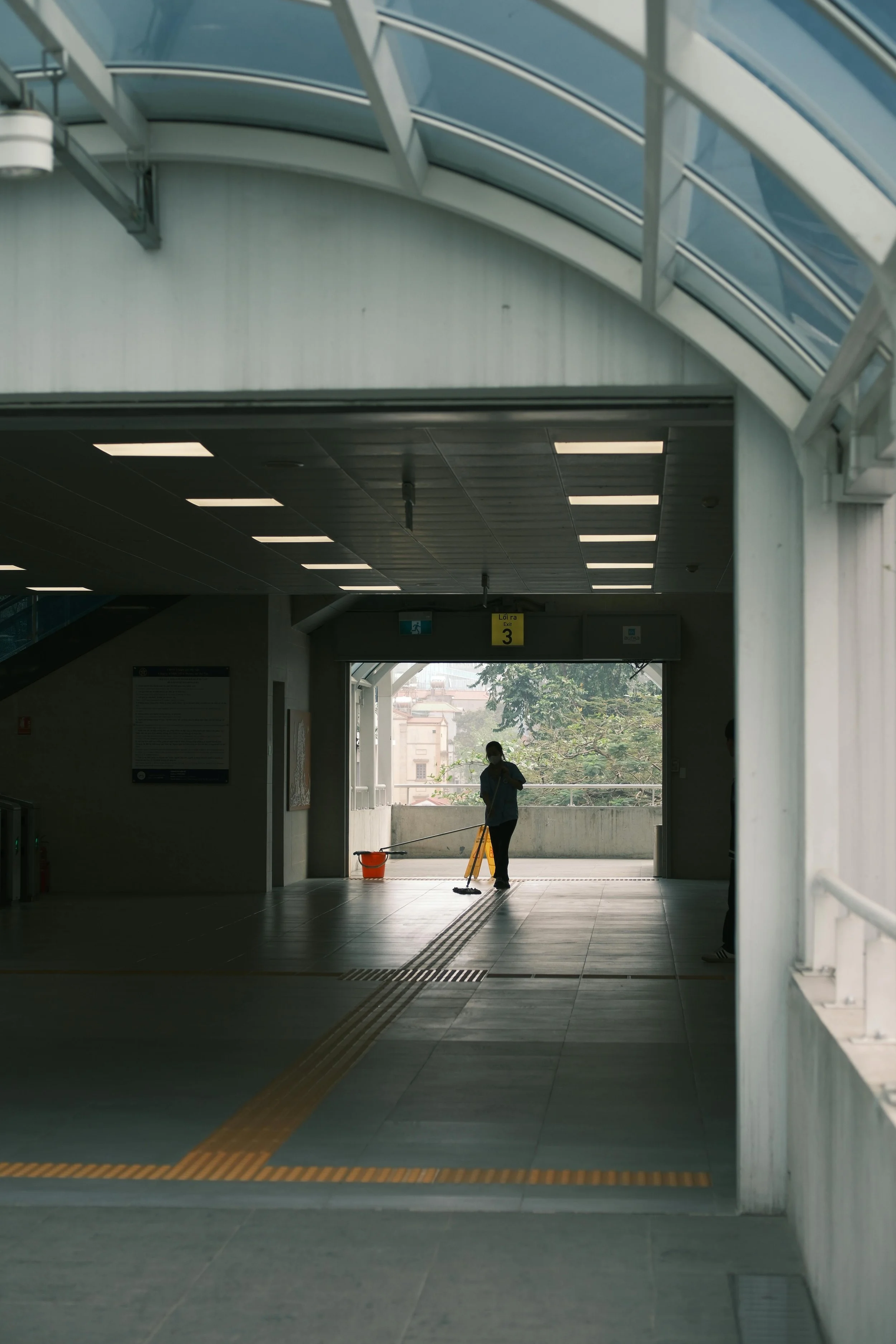 A person cleaning the floor inside a building with a mop, near an open exit door with a yellow caution sign, in a modern structure with a glass ceiling and bright lighting.