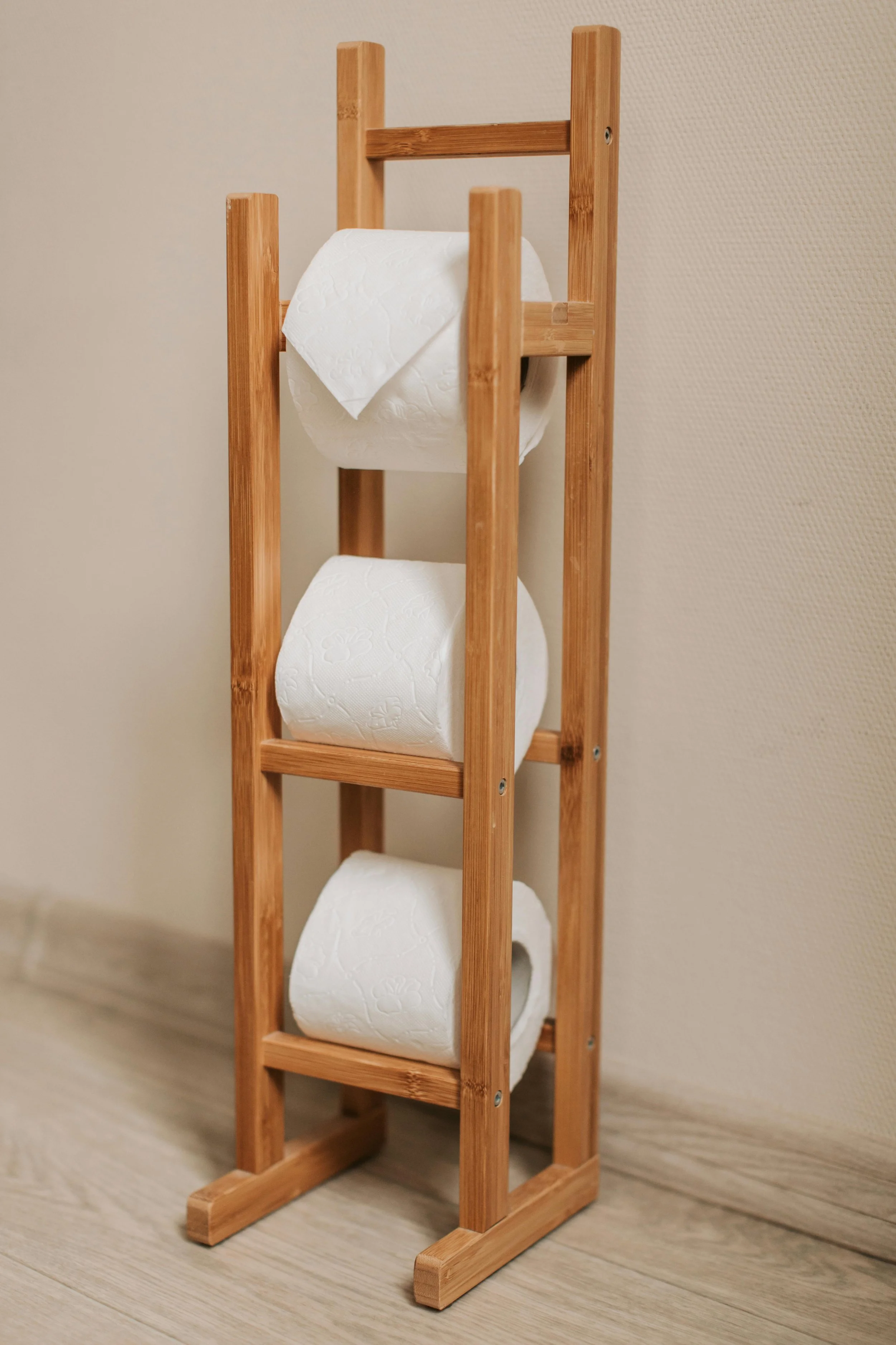 Wooden toilet paper holder with three rolls of white toilet paper, standing on a wooden floor against a beige wall.
