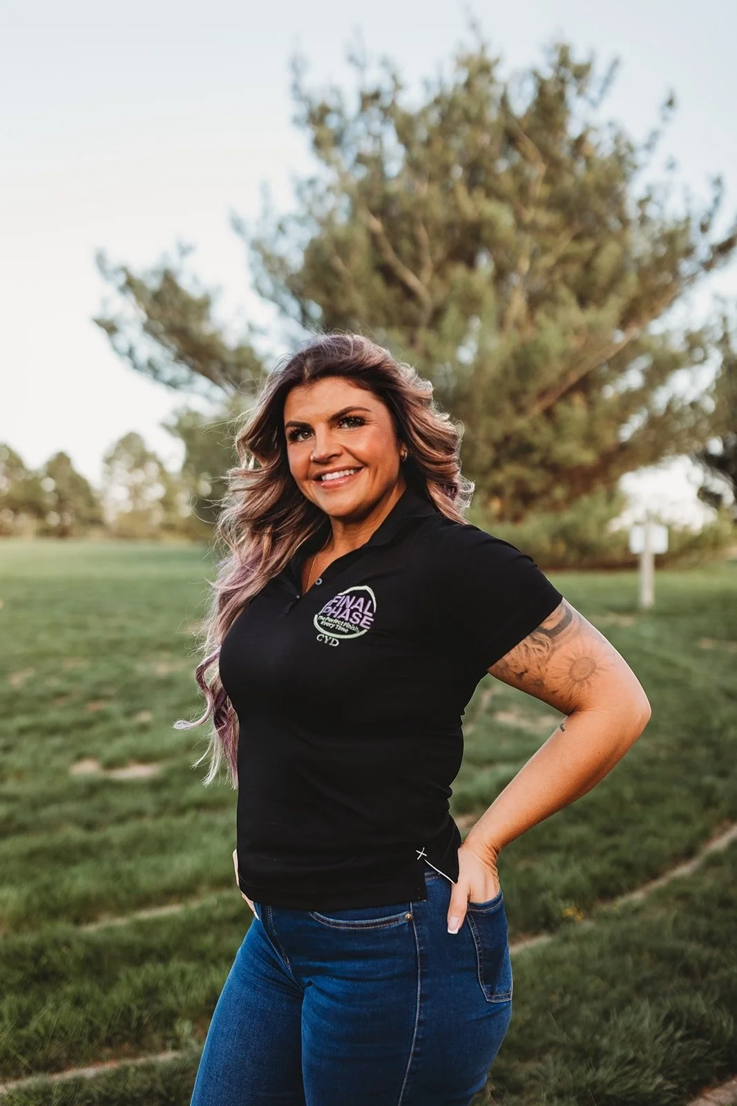 A woman with long, wavy hair and tattoos on her arm, smiling and standing outdoors in front of a tree, wearing a black shirt and blue jeans.