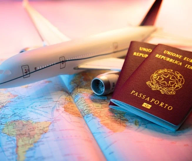 An airplane model, two Italian passports, and an open world map placed on a desk, with a pink background.