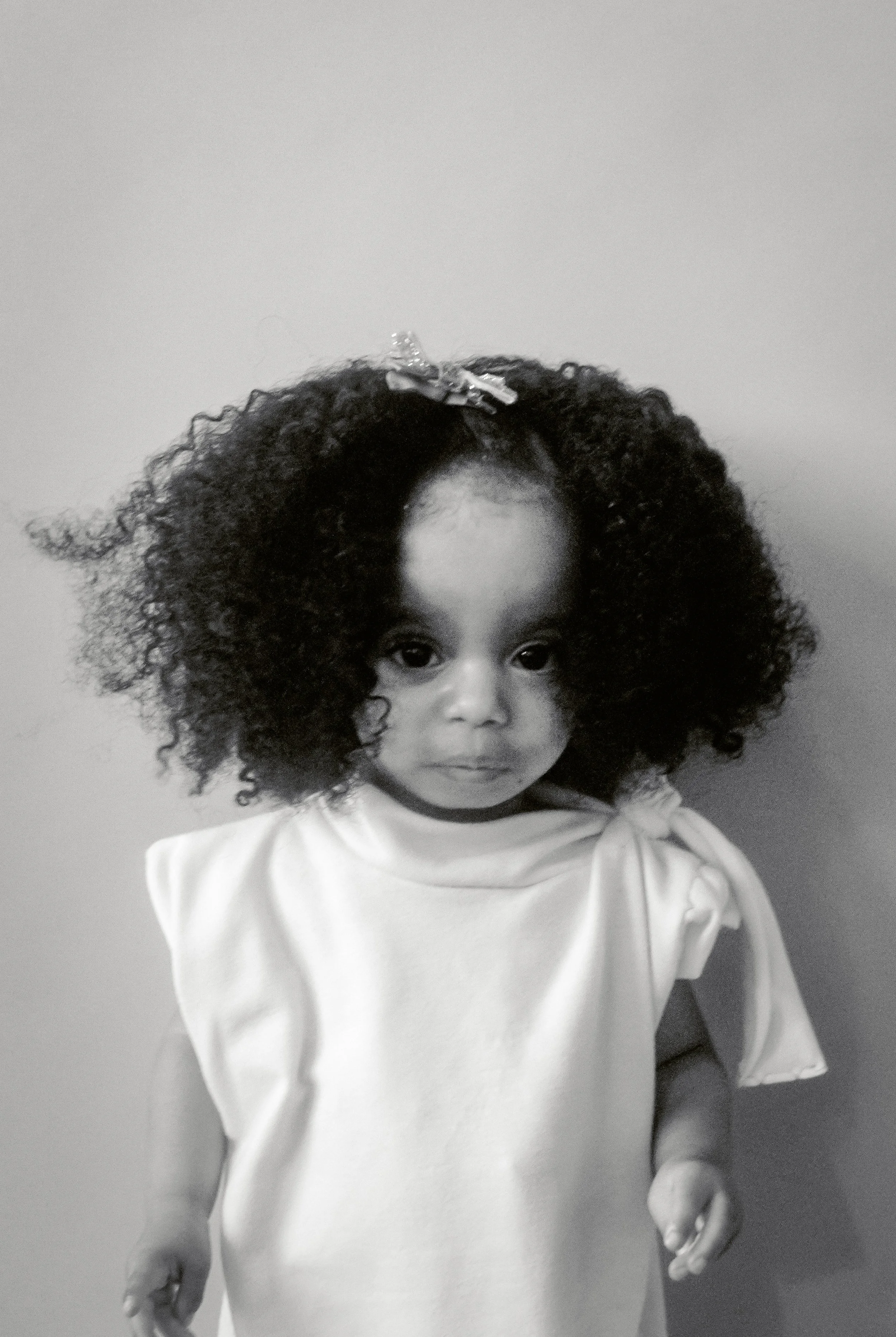 A young girl with curly hair, wearing a light-colored top, standing against a plain wall.