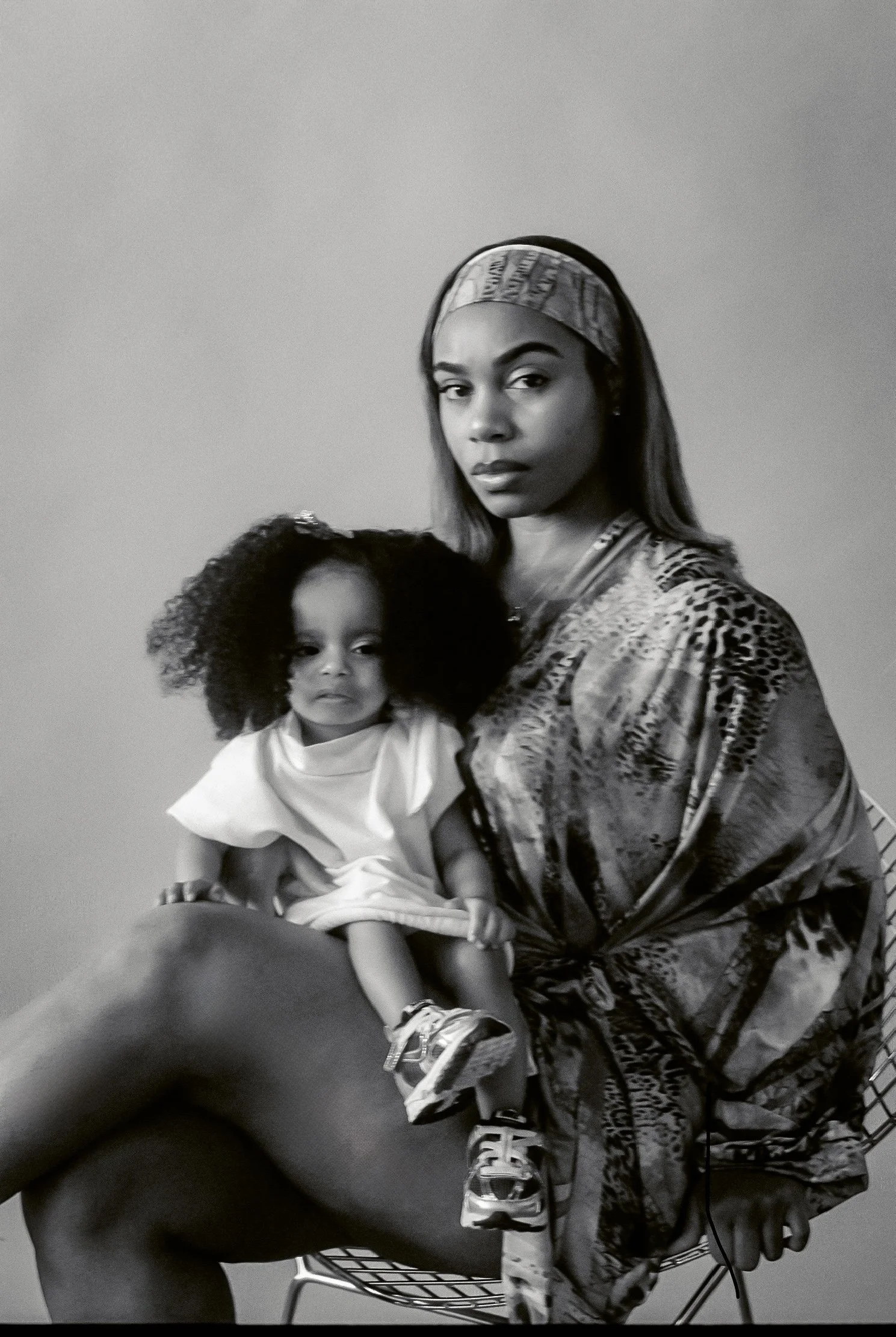 A woman sitting on a shopping cart holding a young girl with curly hair. The woman is wearing a headscarf and animal print clothing, while the girl is dressed in a white outfit and starts at the camera.