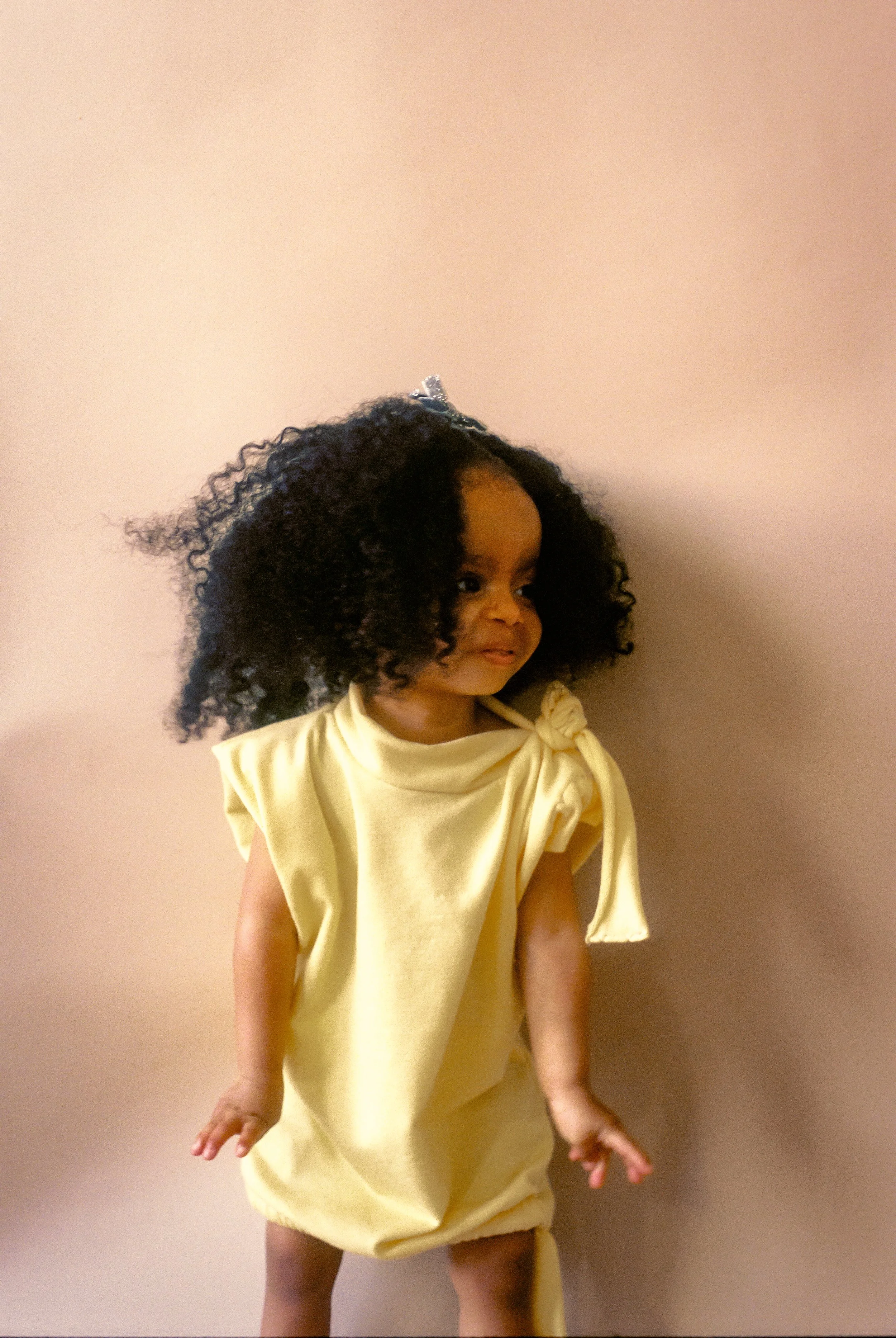 A young girl with curly black hair, wearing a light yellow dress, standing against a light pink wall.