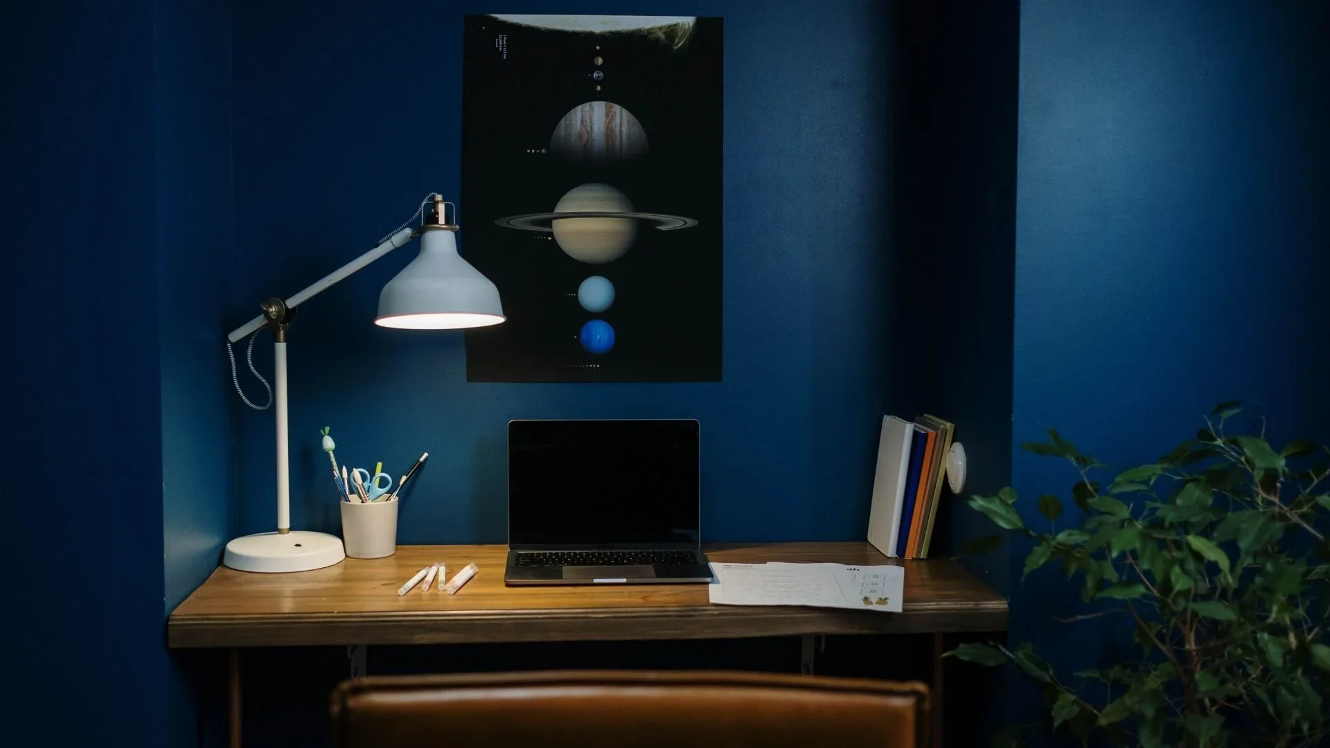 A neatly organized study desk against a deep blue wall featuring a white adjustable desk lamp, a pen holder with pens and scissors, a laptop, notebooks, and some chalks. There is a space-themed poster of planets above the desk. A leafy plant is partially visible on the right side.