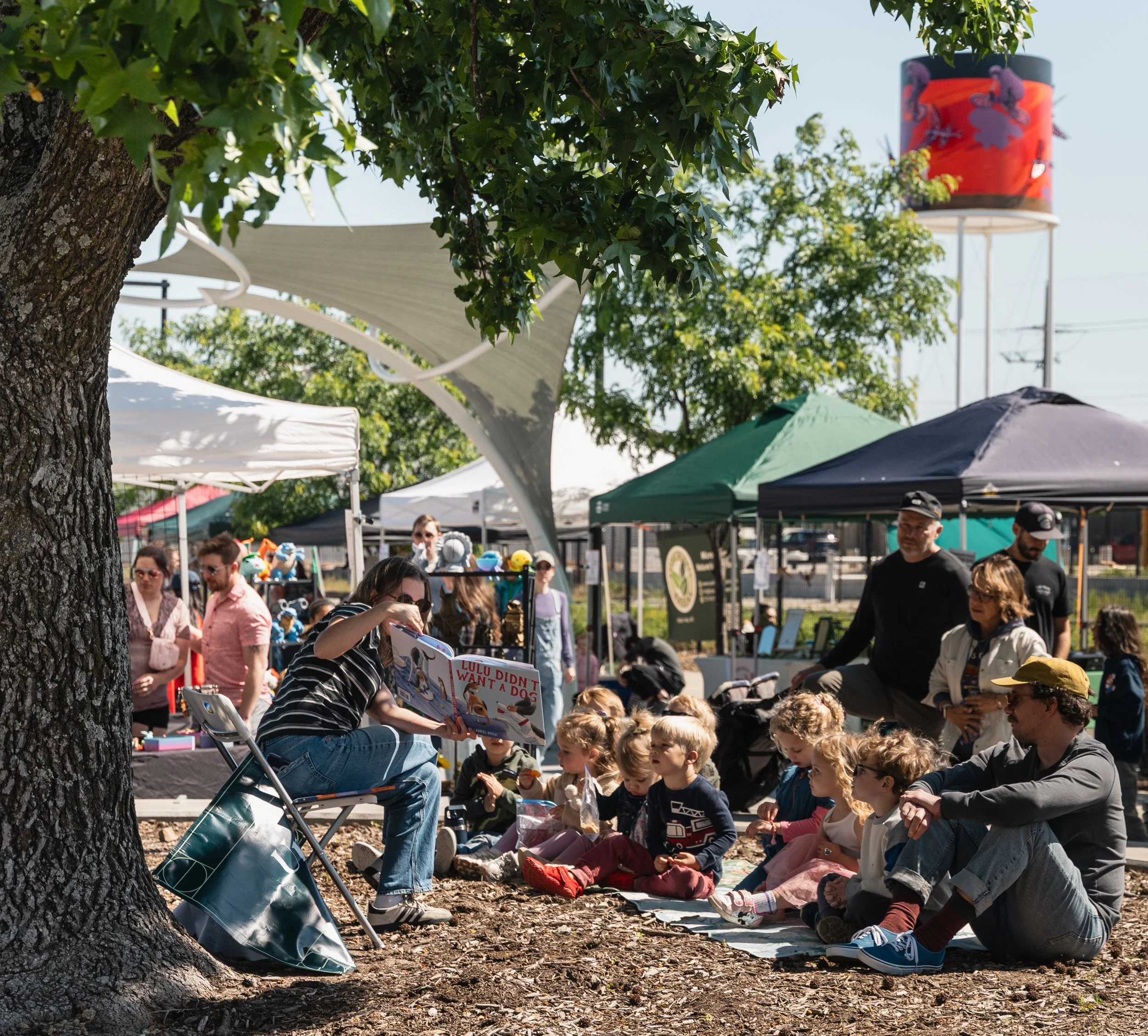 A diverse group of people, including children and adults, gathered outdoors at a daytime event under tents with trees in the background. A woman is seated reading a colorful book to an attentive crowd of children seated on the ground, and a man is standing nearby among other attendees.