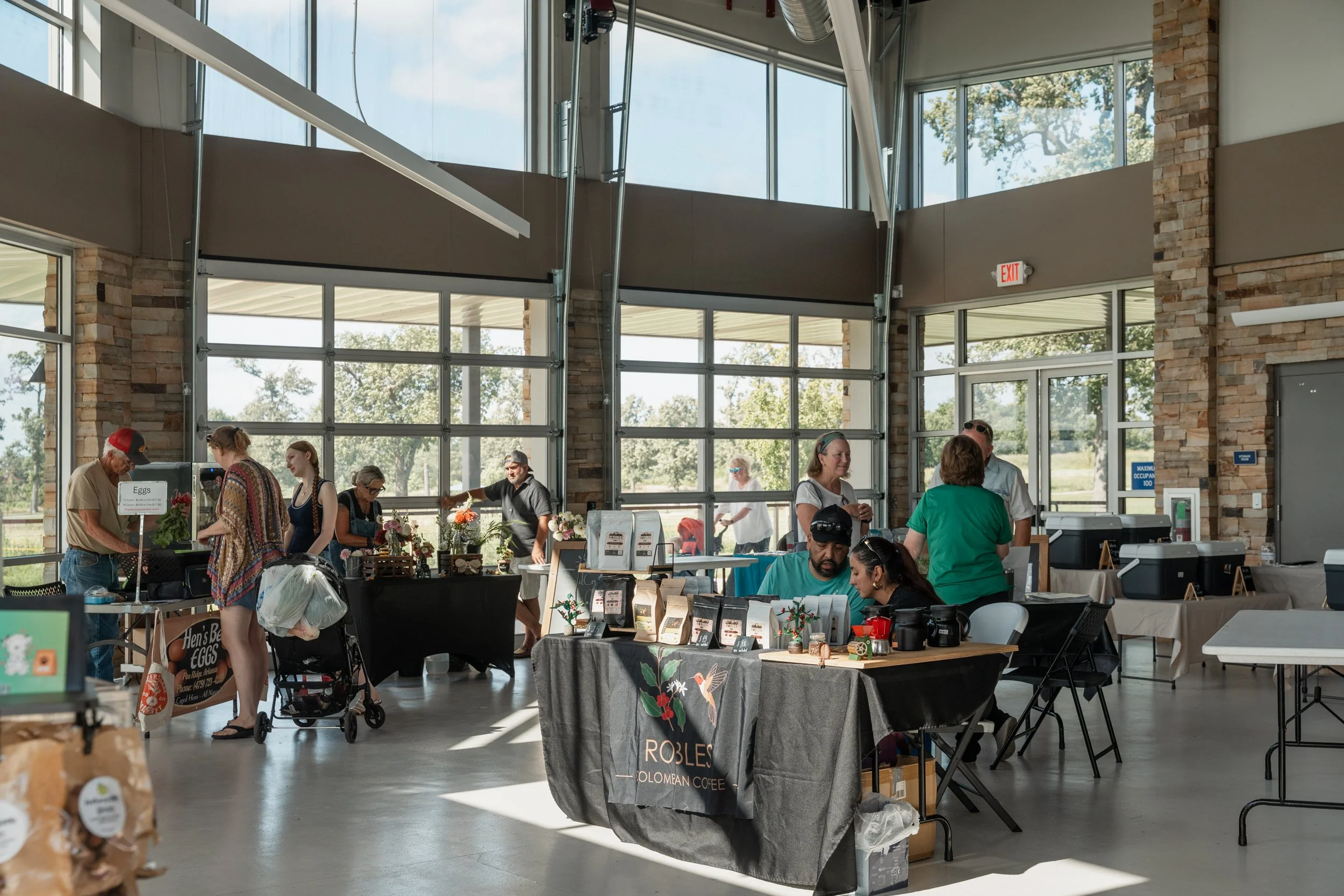 People shopping at an indoor farmer's market or craft fair with tables displaying various products, large windows, and a brick wall in the background.